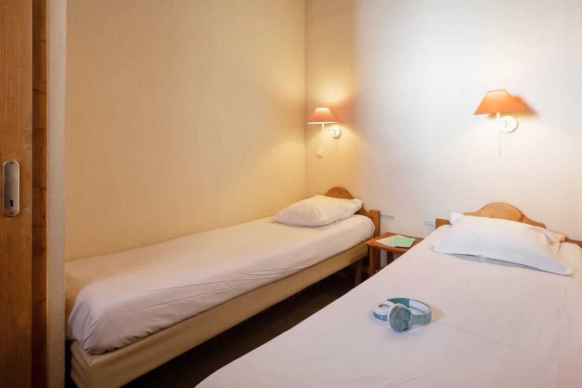 Une petite chambre d’hôtel composée d’une seule pièce, avec deux lits, une table de chevet et deux lampes fixées au mur.