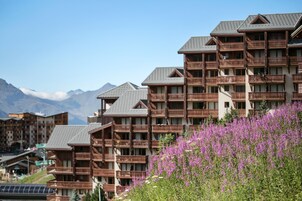 A hotel with multiple balconies, a mountainous backdrop, and a field of purple flowers in the foreground.