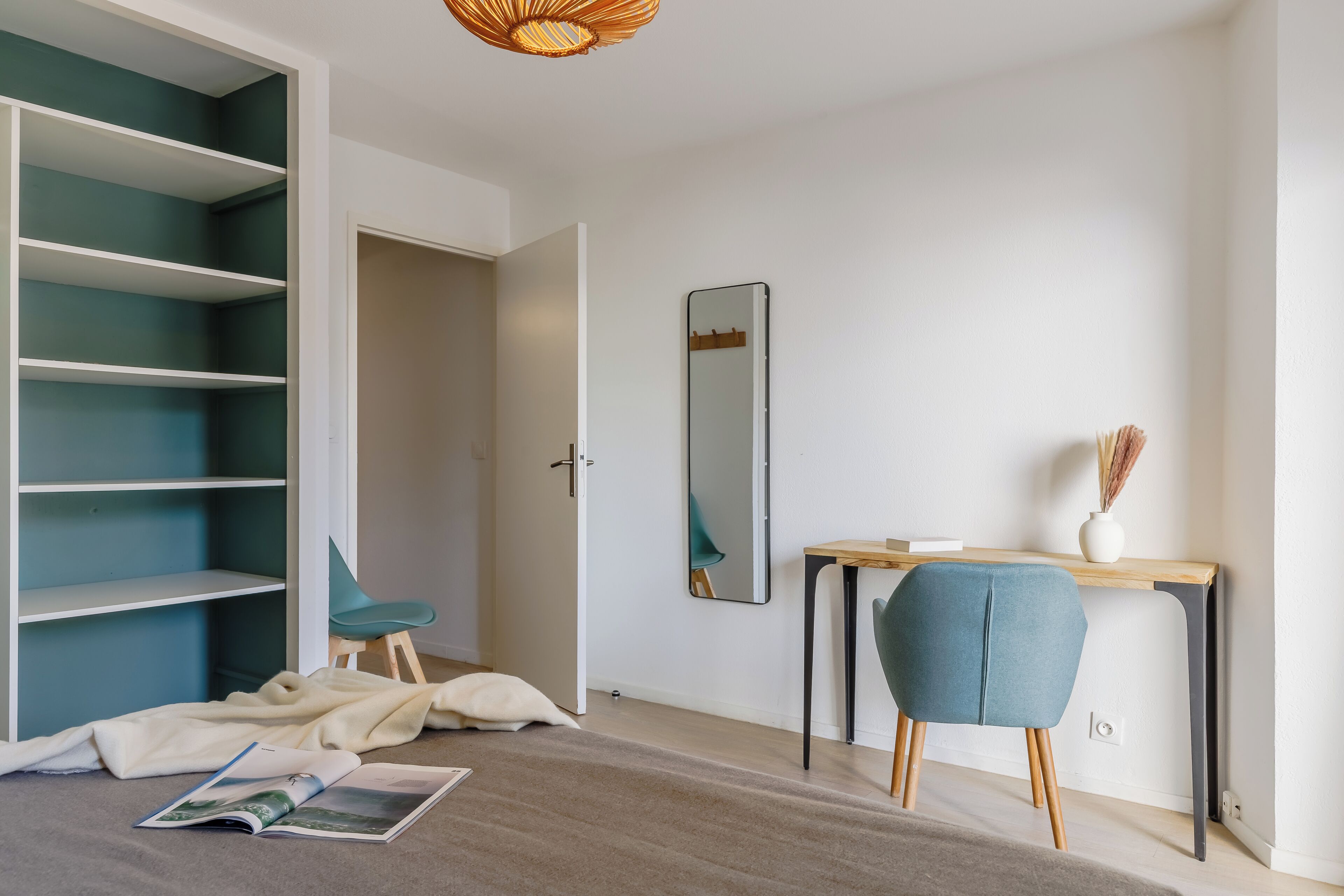 Une pièce comprenant une grande étagère turquoise, un bureau en bois avec une chaise, un miroir et un vase contenant des plantes séchées.