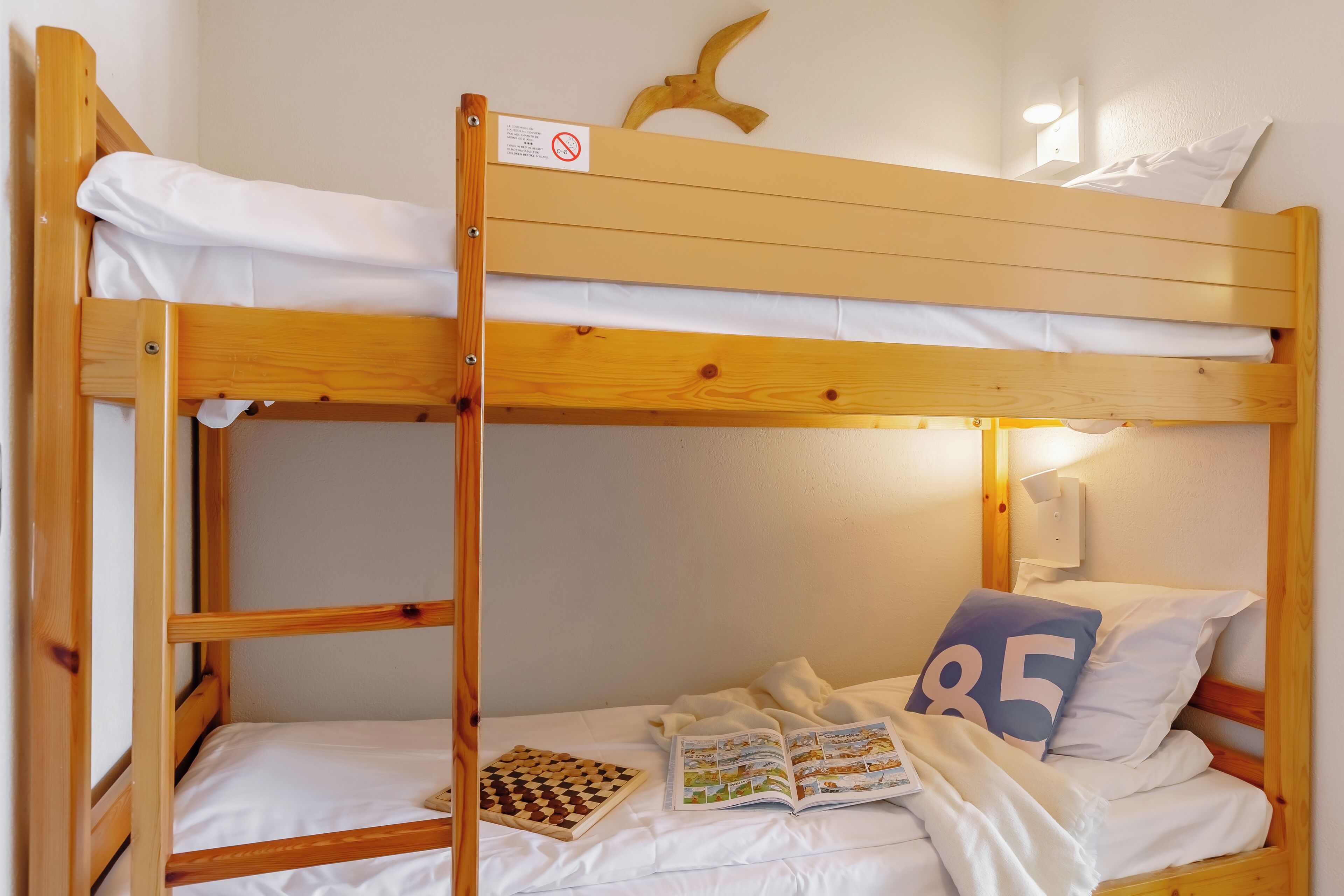 A bunk bed with white bedding, a wooden headboard, a wall-mounted light, and a number 85 pillow.