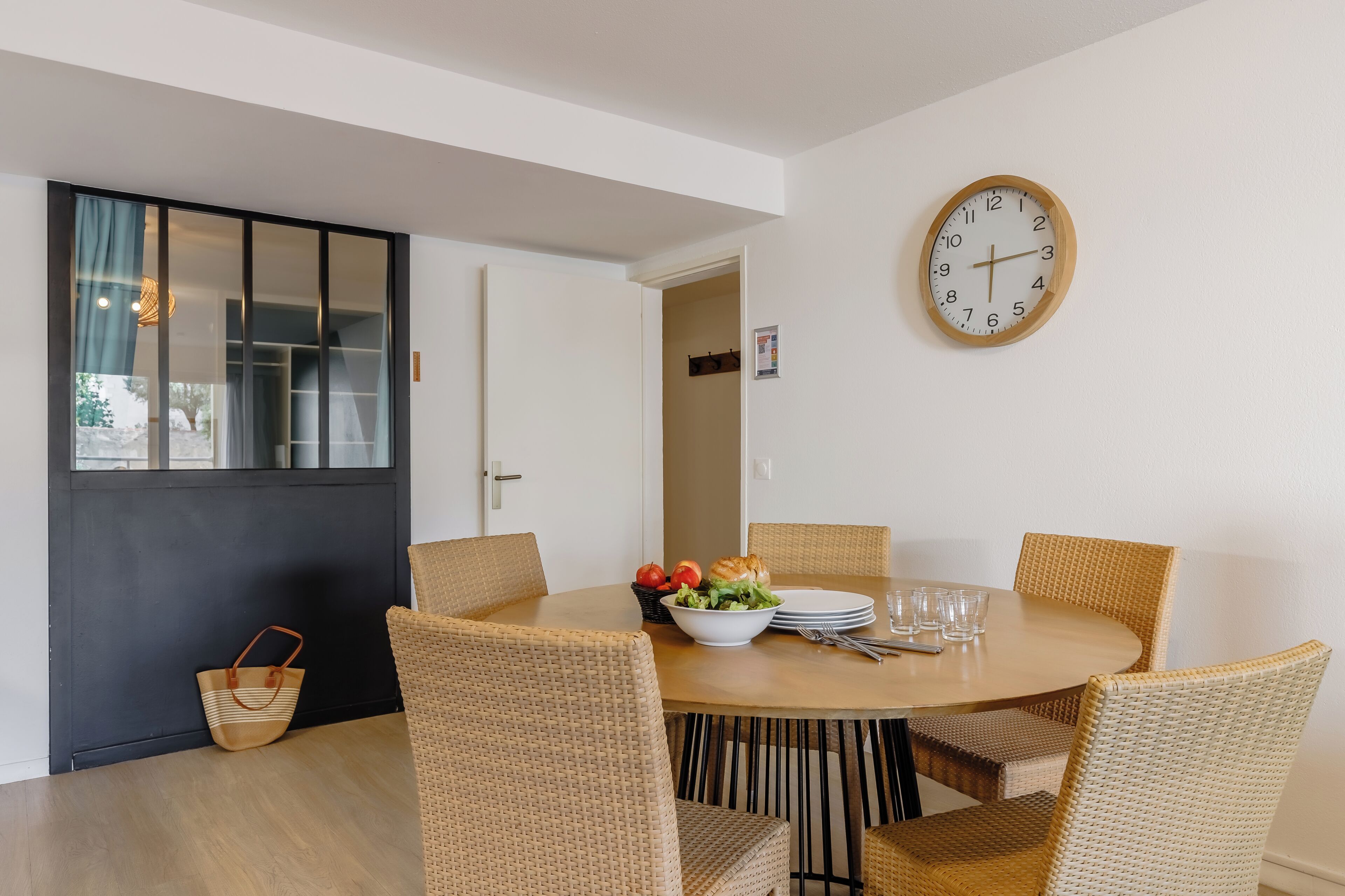 A dining area with a round table, wicker chairs, a bowl of salad, and a clock on the wall.