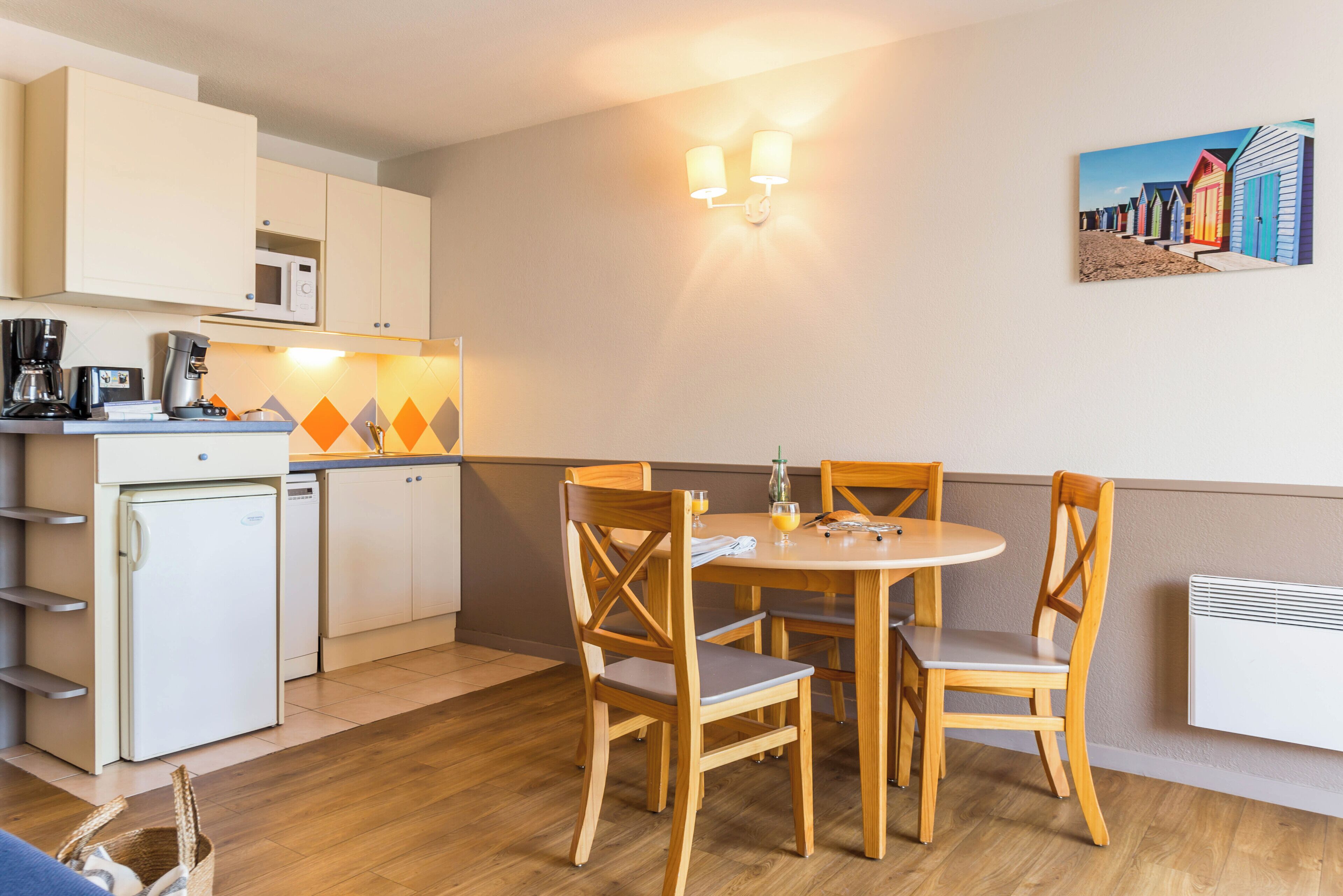 Una cocina compacta con refrigerador, microondas y cafetera, un área de comedor con una mesa redonda y sillas de madera, y un aplique de pared.