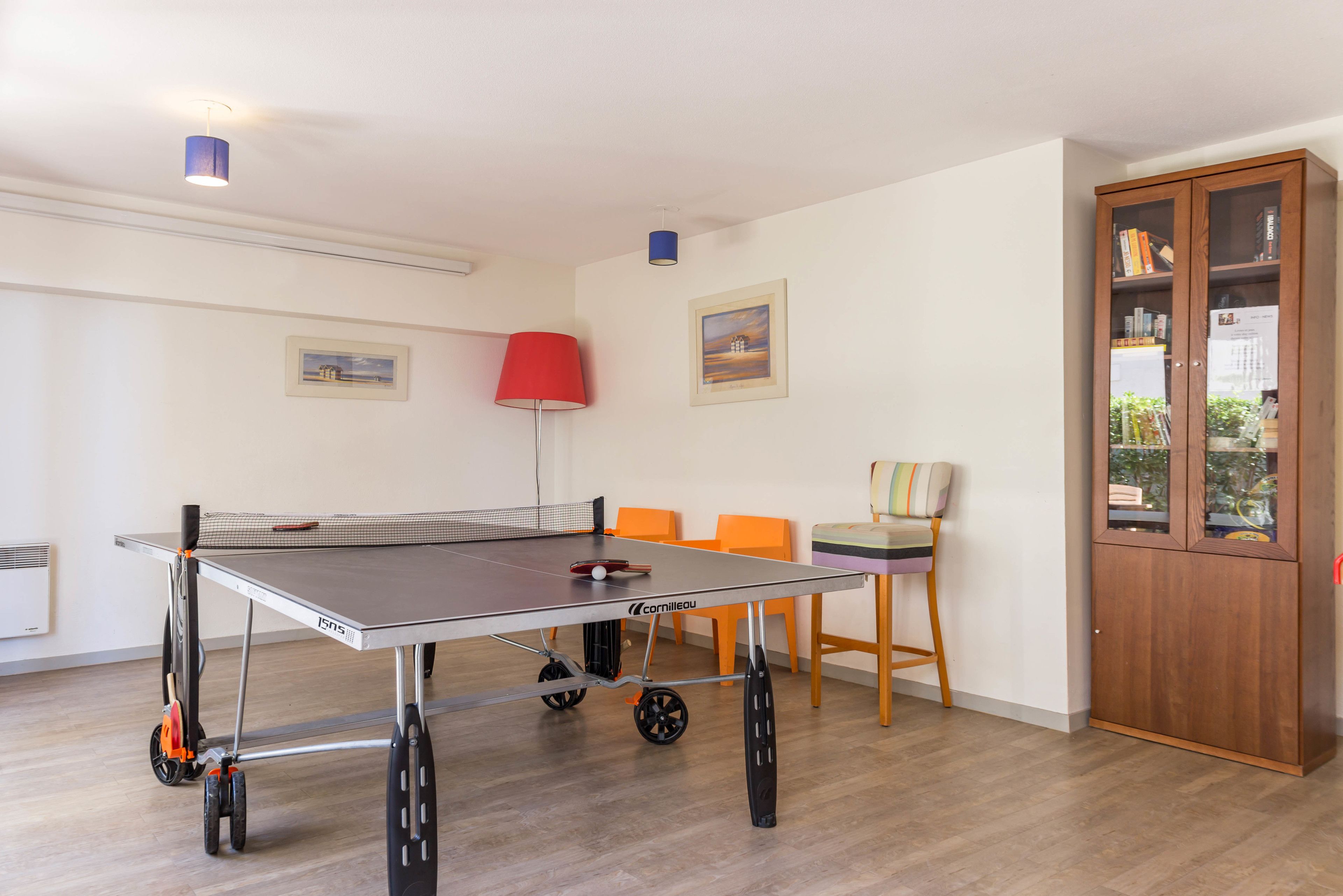 A ping pong table with wheels, orange chairs, a red lamp, and a wooden cabinet with books.