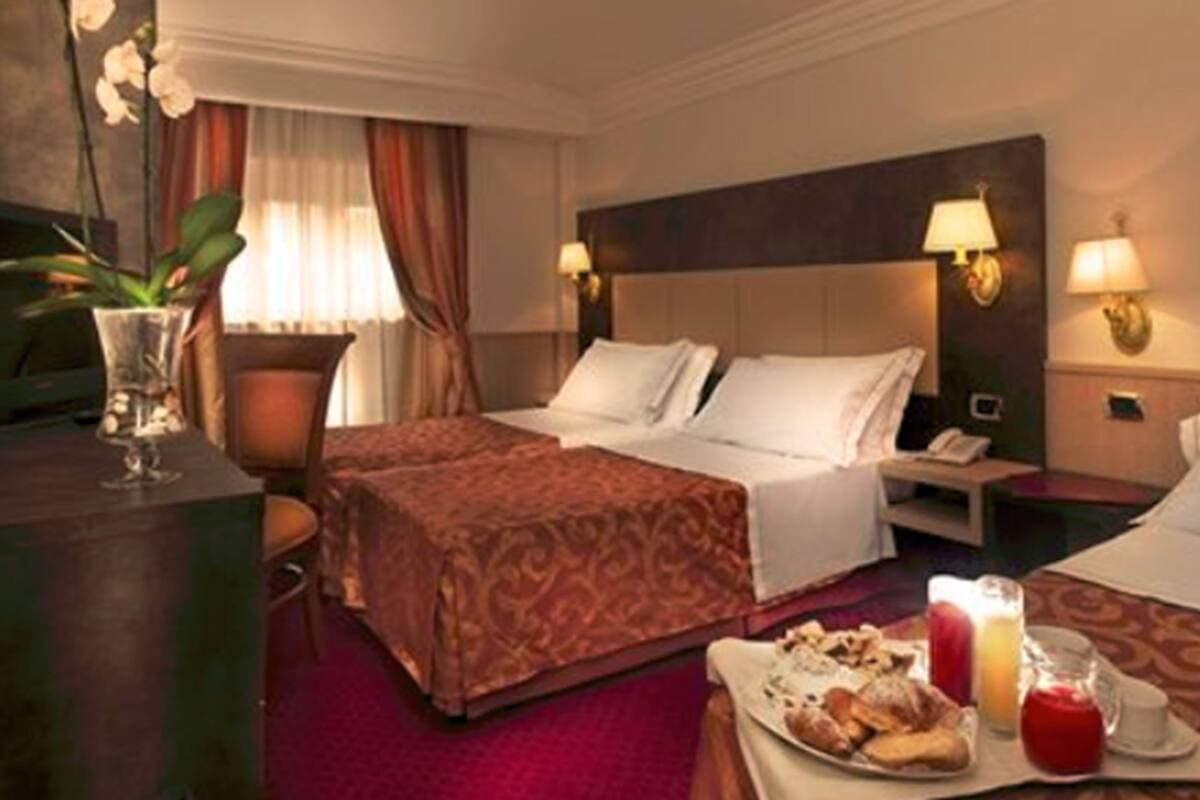 A hotel room with two beds, a desk, a chair, a TV, and a dining table with breakfast items.