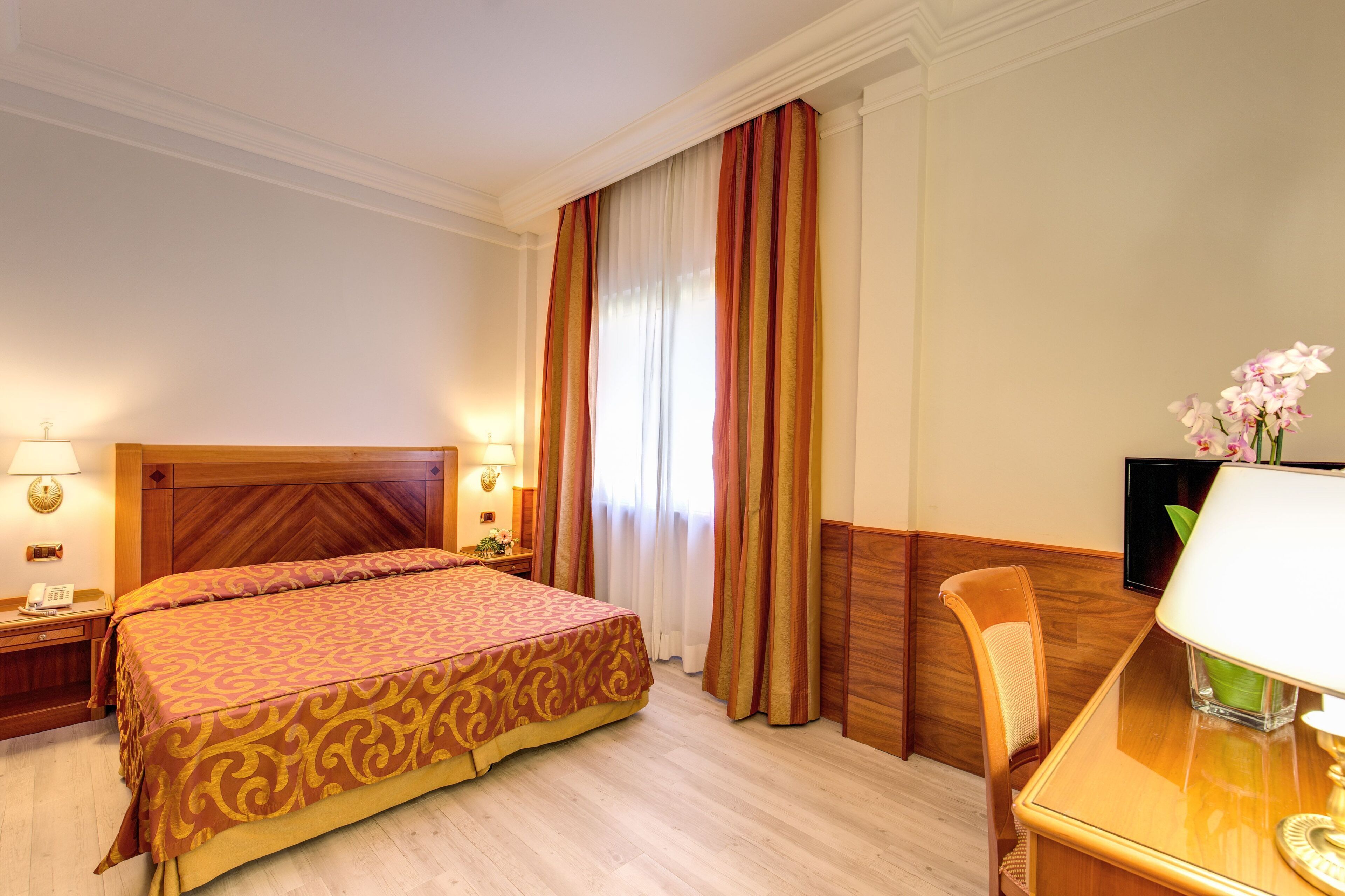 A hotel room with a large bed, wooden headboard, two bedside lamps, a desk with a chair, and a TV.