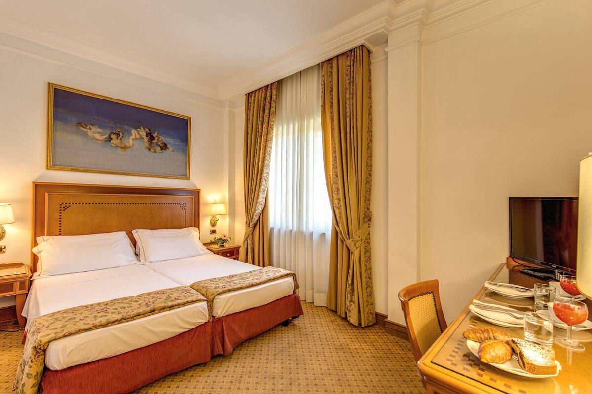 A hotel room with a double bed, a television, a dining table with breakfast items, and a painting on the wall.