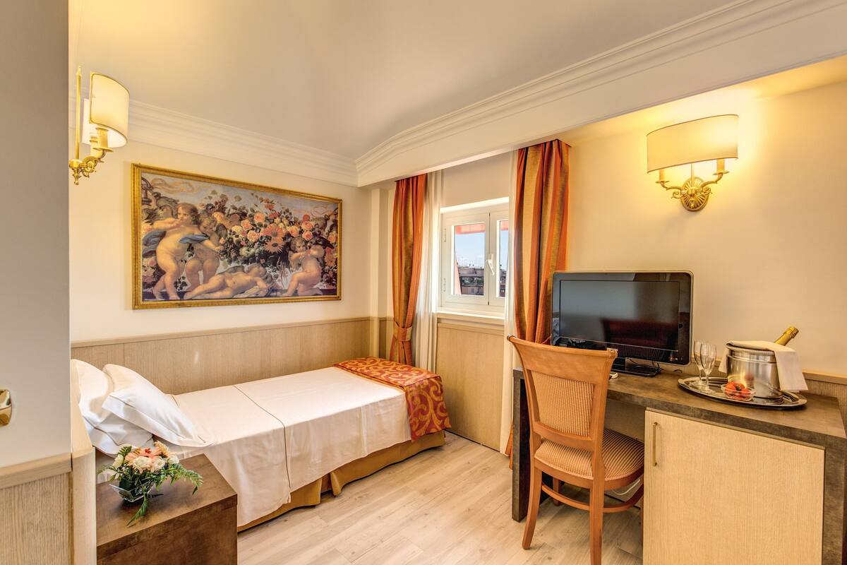 A hotel room with a bed, a desk with a TV, a chair, a window with curtains, and a painting on the wall.