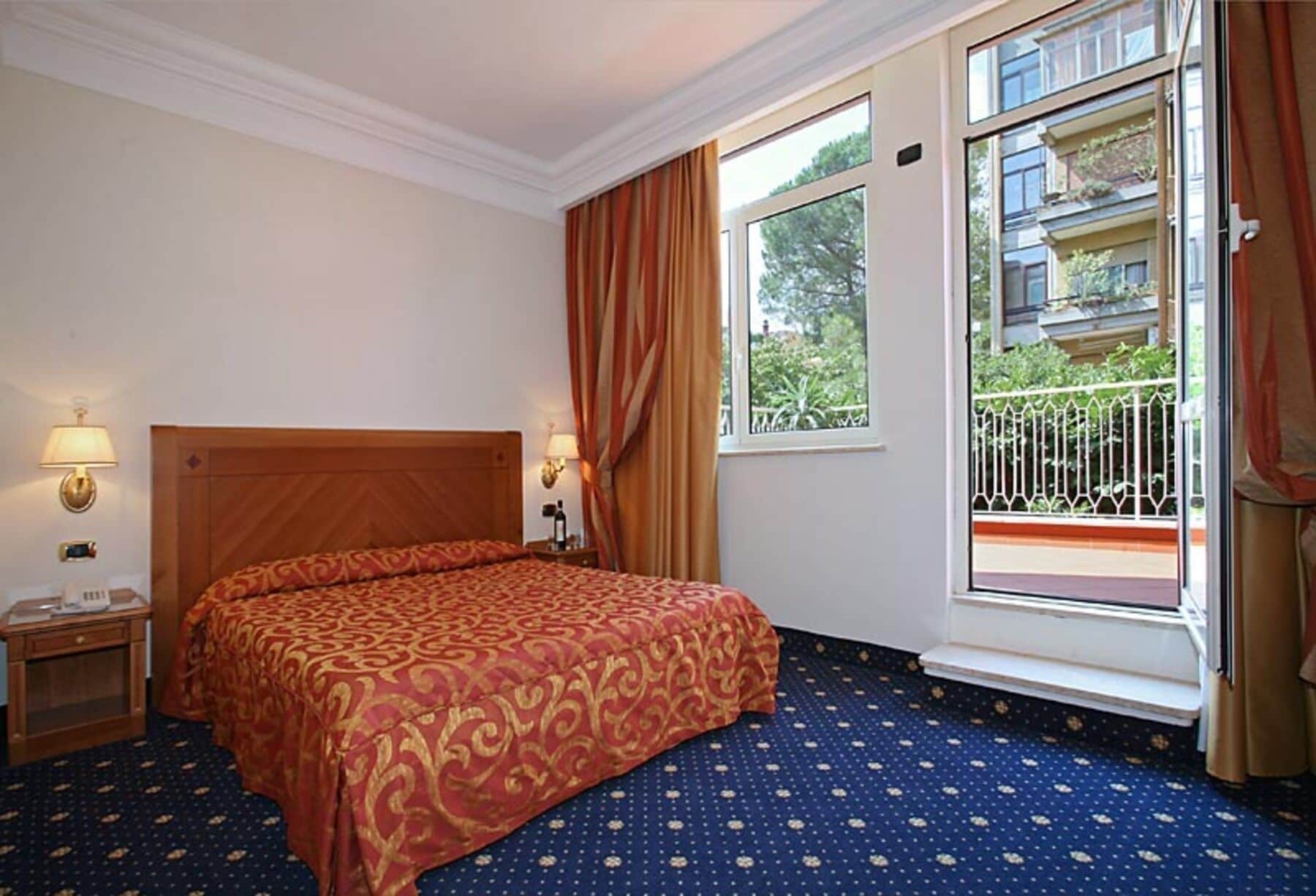 A hotel room with a large bed, a nightstand, a lamp, a window with curtains, and a balcony with a railing.