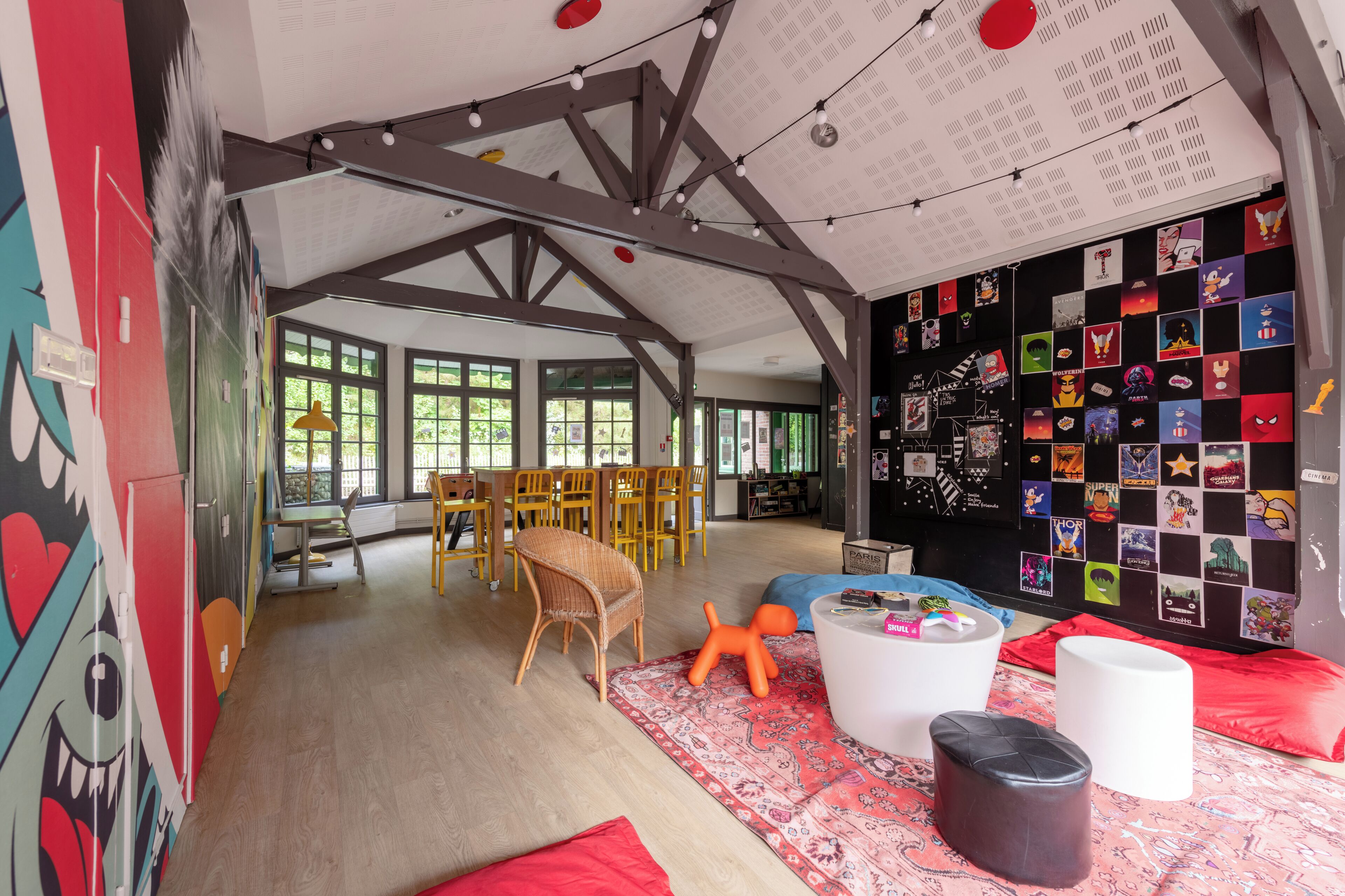 Une pièce spacieuse avec un plafond haut, des poutres en bois et une grande fenêtre. On y trouve un coin repas avec des chaises et une table, ainsi qu’un mur décoré de divers autocollants.