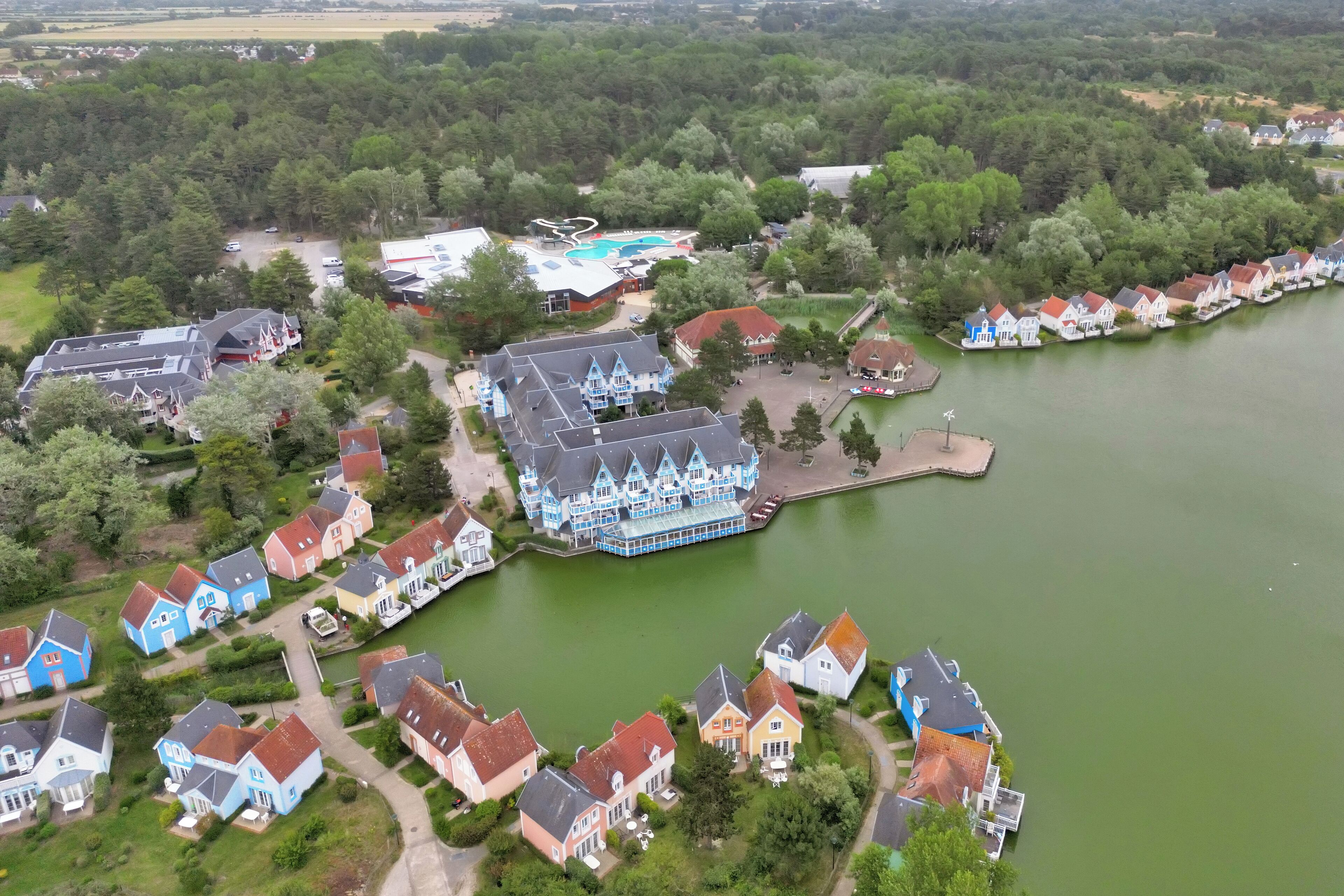 Vista aérea de una zona residencial junto a un lago, con casas, un edificio grande azul y una piscina.