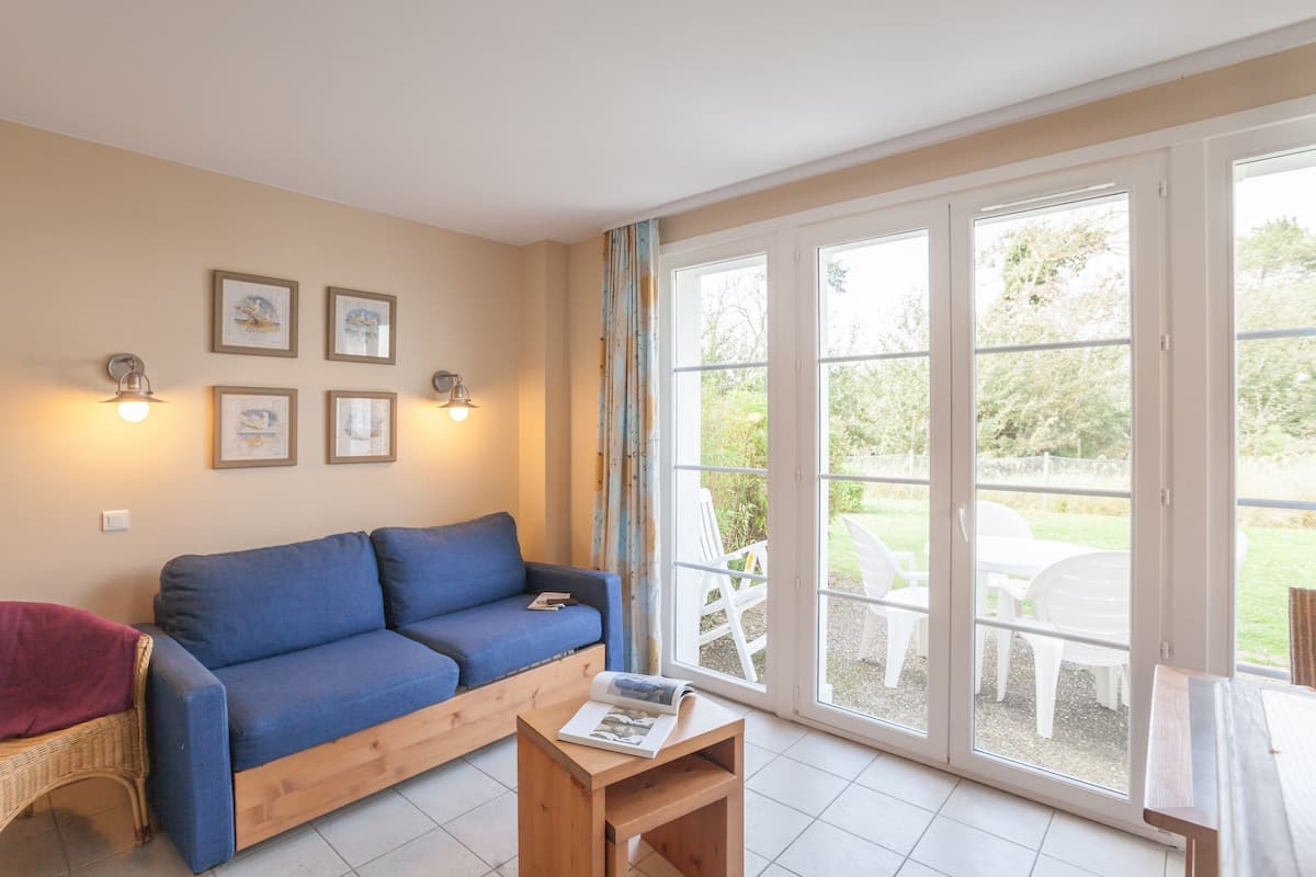Un salon avec un canapé bleu, une table basse en bois sur laquelle repose un magazine, et un fauteuil en osier. Des tableaux encadrés ornent le mur, et une porte-fenêtre coulissante donne accès à un espace extérieur où se trouvent des chaises blanches.