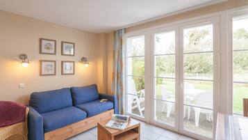 Een woonkamer met een blauwe bank, een houten salontafel met een tijdschrift, en een rieten stoel. Er hangen ingelijste foto’s aan de muur en een schuifdeur leidt naar een buitenruimte met witte stoelen.