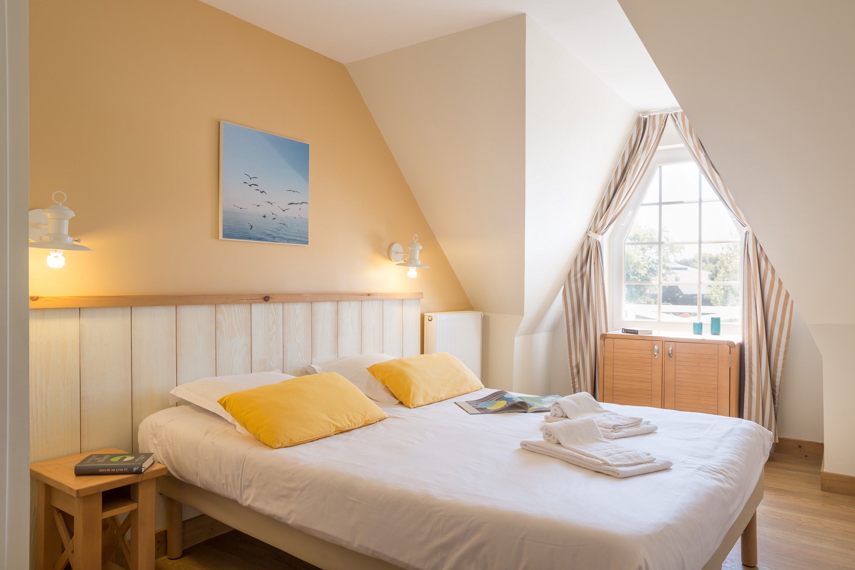 A bedroom with a bed, pillows, a bedside table, a lamp, a window with curtains, and a painting on the wall.