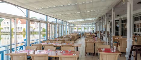Un área de comedor al aire libre techada con mesas y sillas, ventanas amplias y vistas a edificios a través de un cuerpo de agua.
