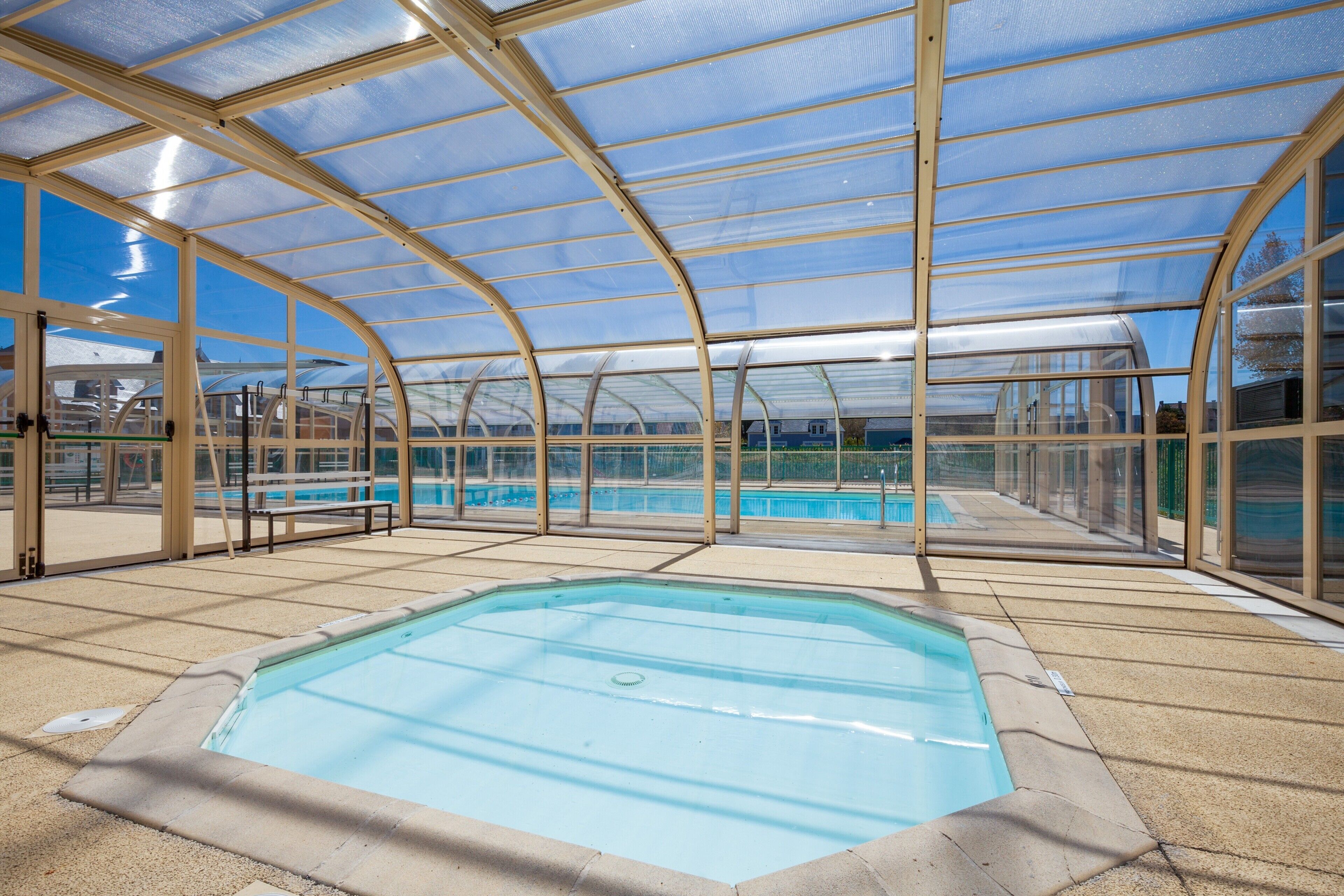 A covered swimming pool with a glass roof, surrounded by a pool deck and benches.