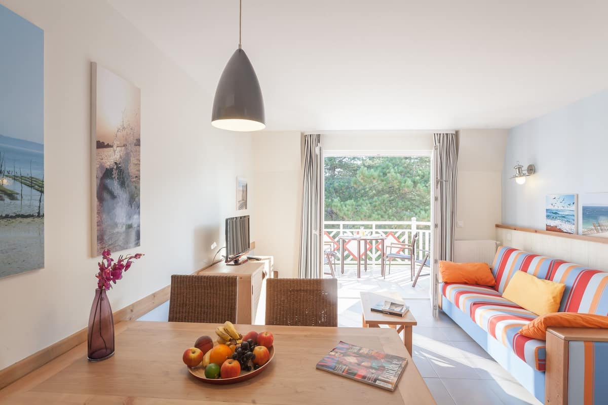 Un salon moderne avec une table à manger en bois, un panier de fruits, un canapé avec des coussins à rayures et un balcon offrant une vue sur la verdure.