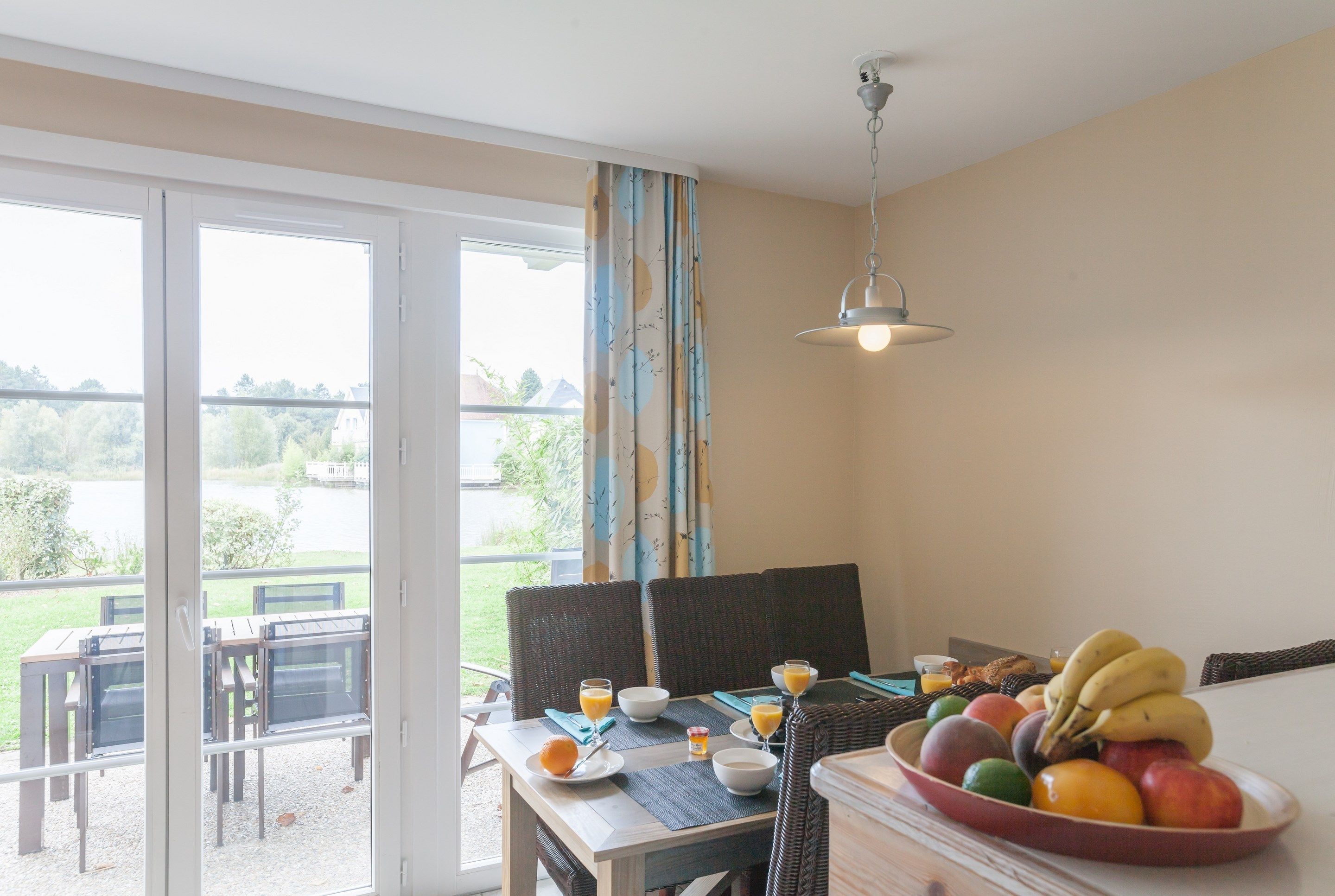 Comedor con una mesa dispuesta para el desayuno, que incluye frutas y vasos de jugo, con vista a un jardín.
