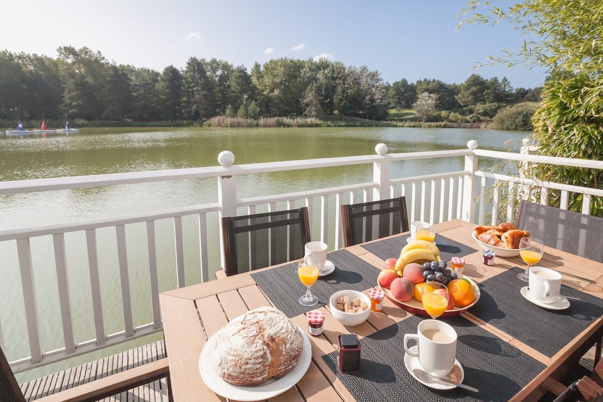 Une table dressée pour le petit-déjeuner, avec du pain, des fruits et des boissons, donnant sur un lac où l’on aperçoit des bateaux au loin.