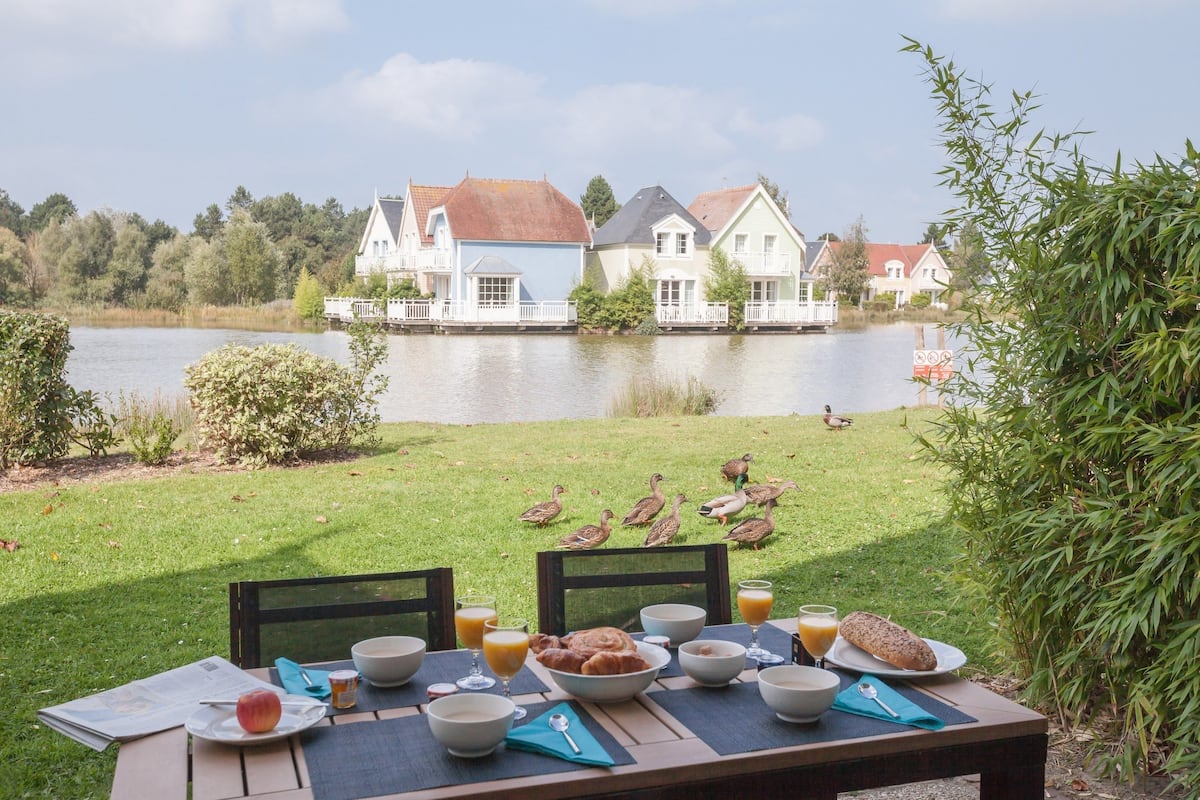 Une table de petit-déjeuner installée au bord du lac, avec des aliments pour le petit-déjeuner, un journal et une vue sur les maisons de l’autre côté de l’eau.