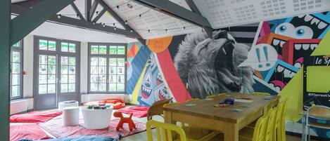 Une pièce avec un plafond haut, de grandes fenêtres et une fresque murale colorée. On y trouve une table en bois avec des chaises, un tapis rouge et un pouf blanc.