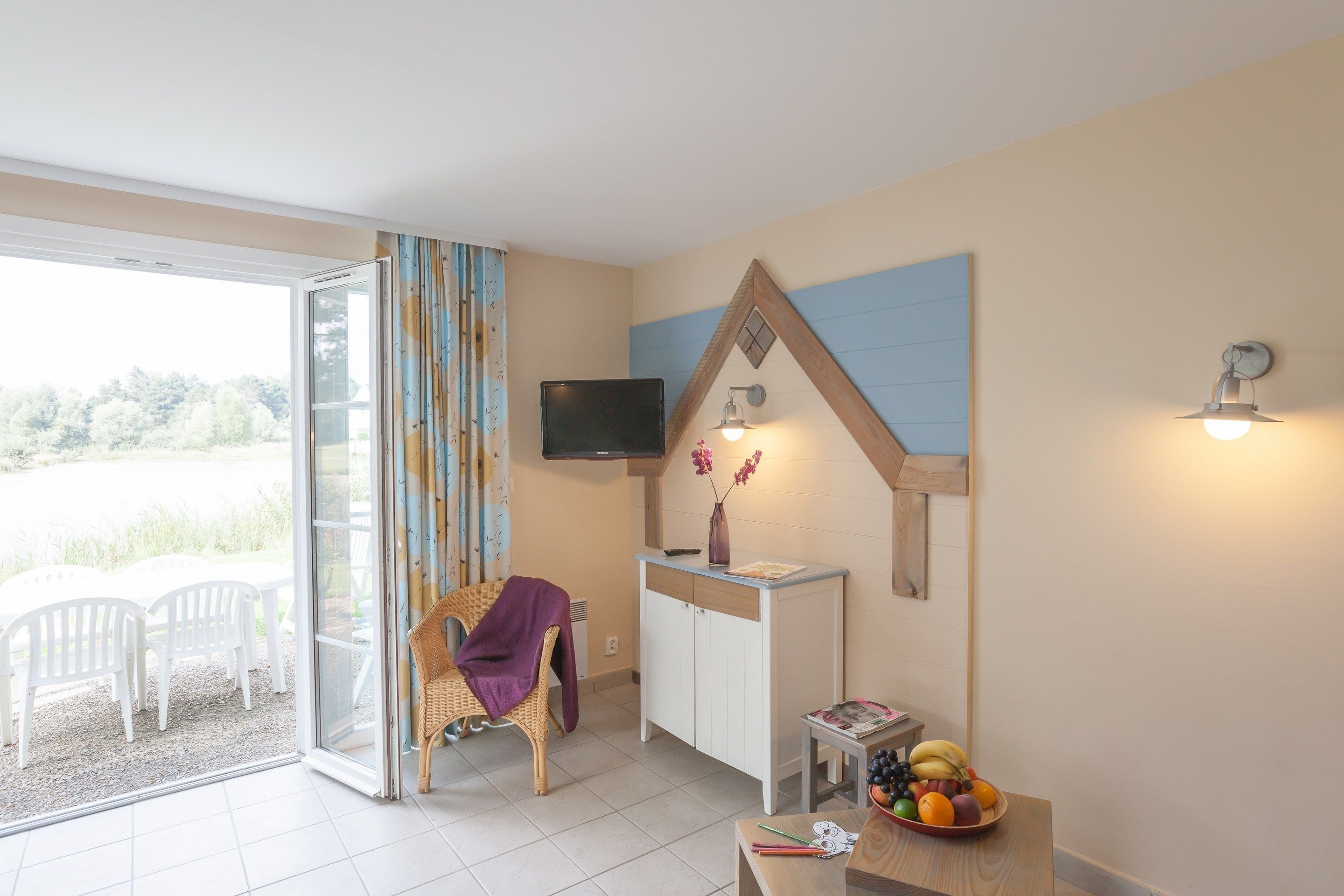 A room with a sliding glass door leading to an outdoor area, a television mounted on the wall, a small table with a fruit bowl, and a wicker chair with a purple throw.