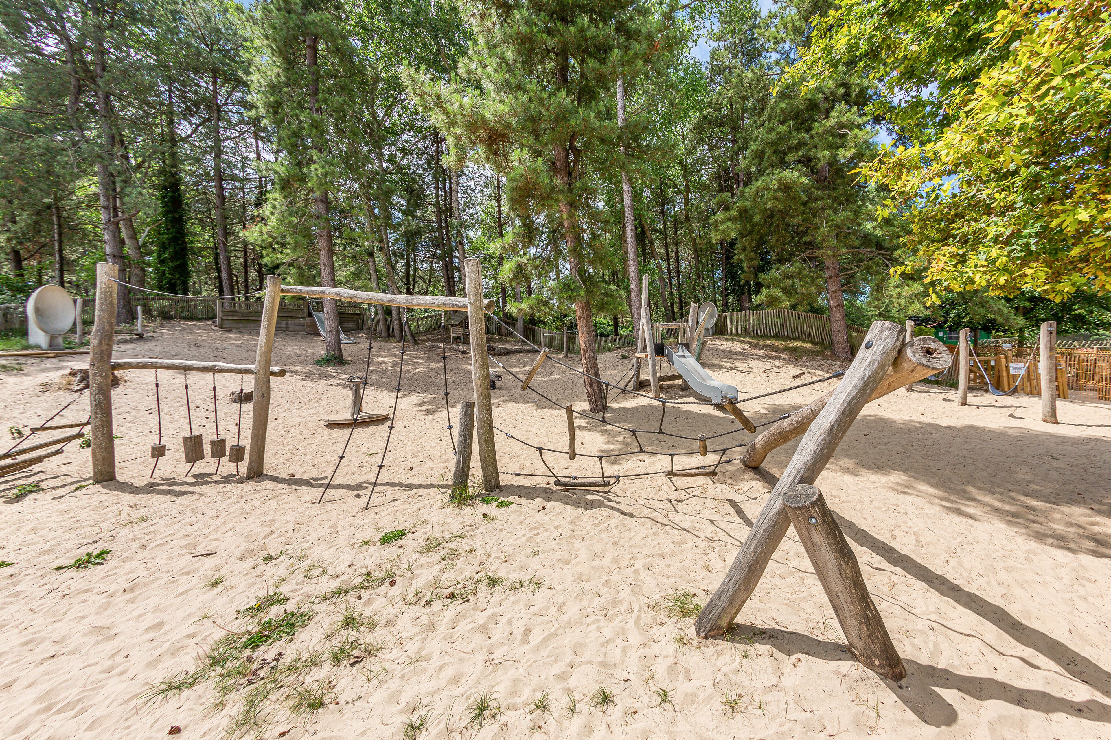 Un terrain de jeux avec des balançoires, des toboggans et des structures d’escalade, le tout entouré d’arbres.