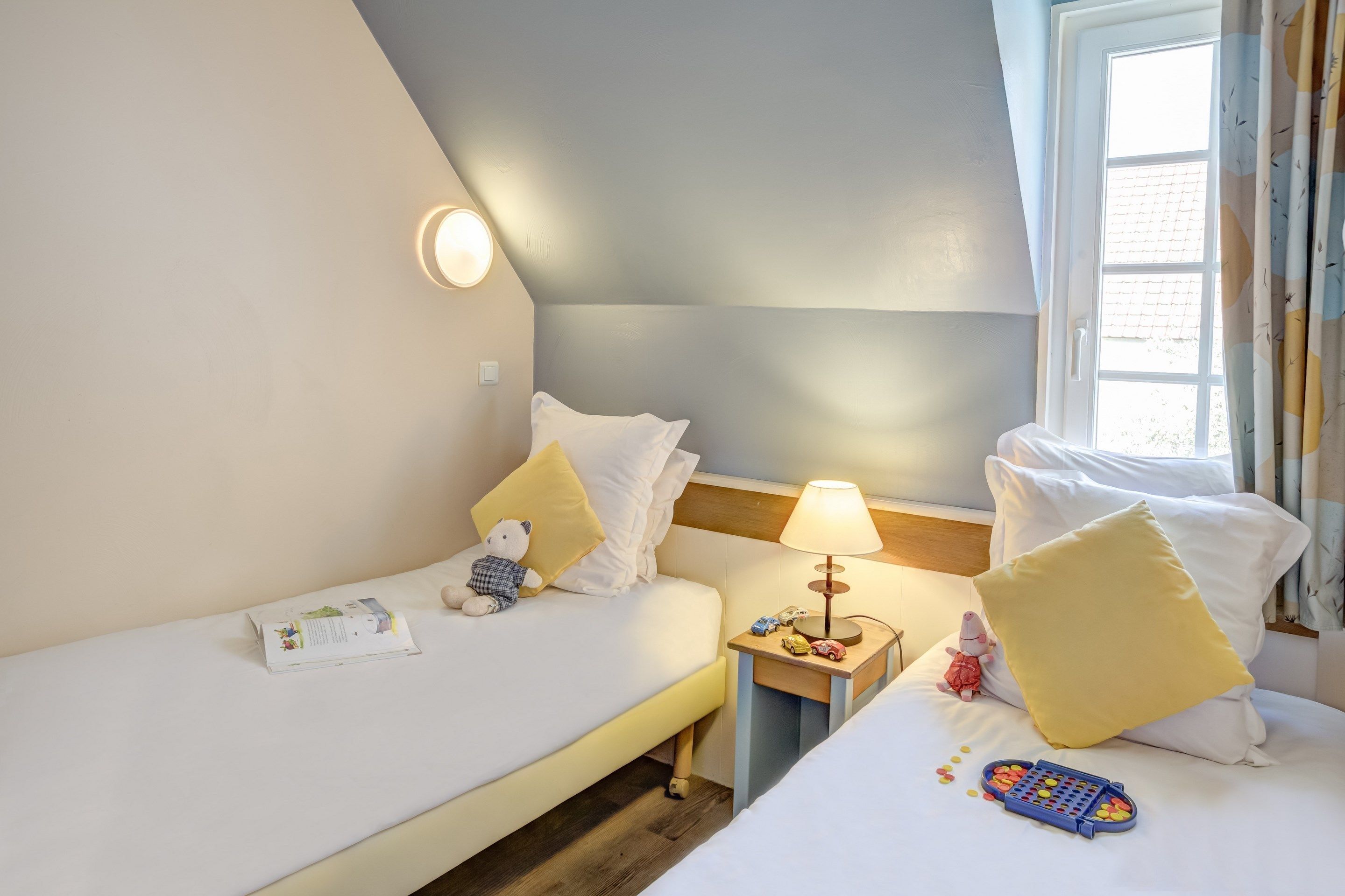 Une chambre d’hôtel avec deux lits, une table de chevet avec une lampe, un ours en peluche et une télécommande.
