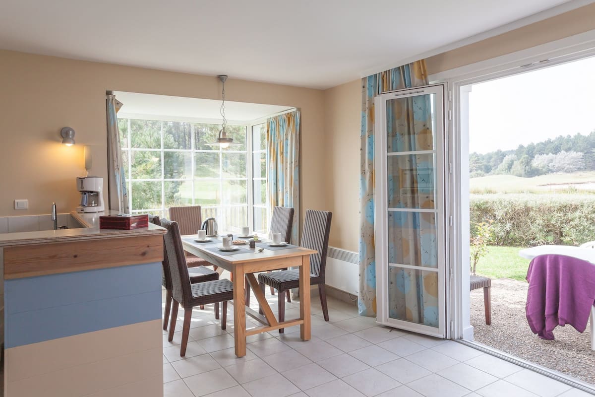 Un espace repas comprenant une table et des chaises, un comptoir de cuisine et une porte-fenêtre coulissante donnant sur un coin salon extérieur.