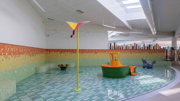 En indendørs swimmingpool med fliset gulv, et gult flag på en stang, og et legeområde med en båd og en hestestruktur.
