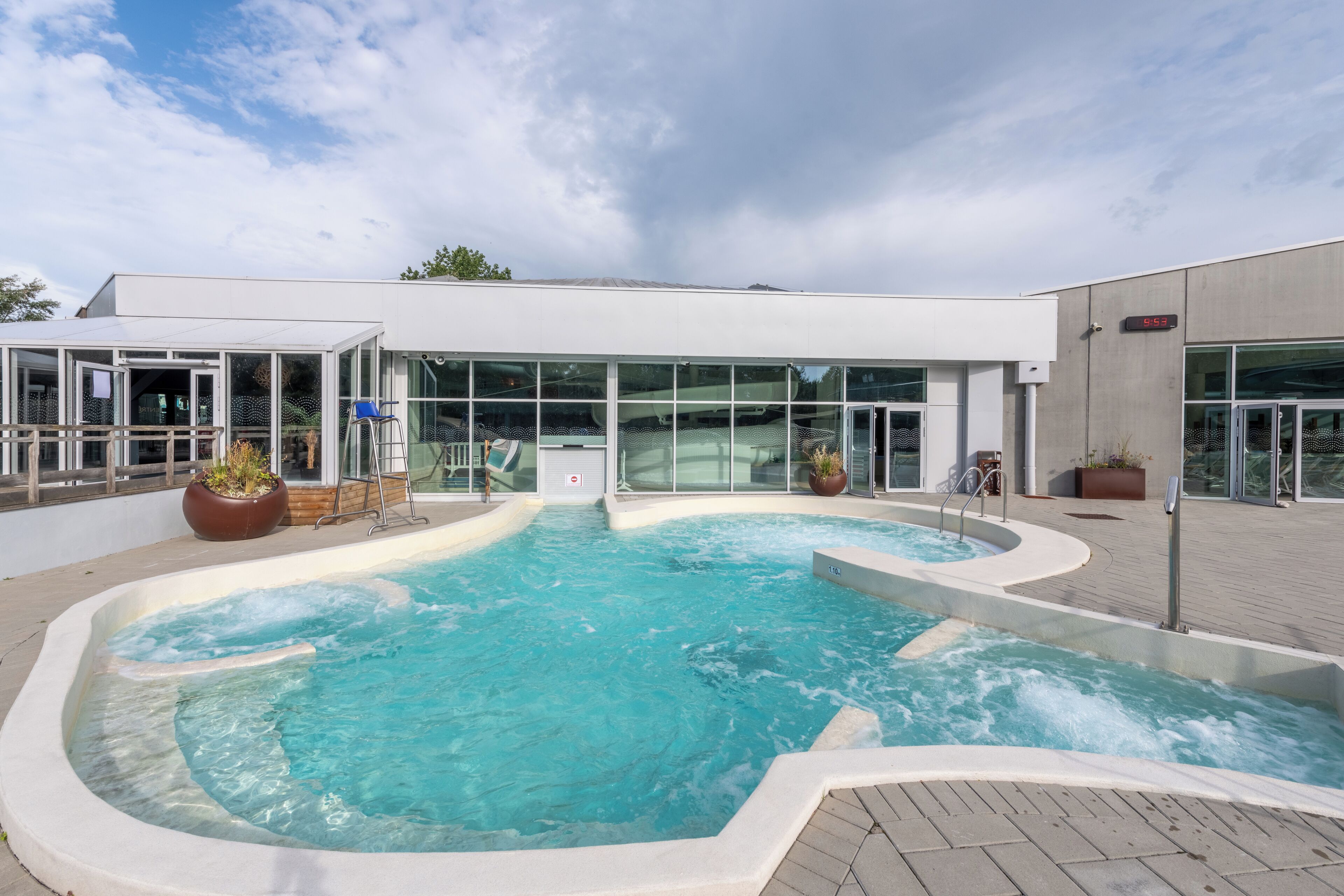 A modern building with a large outdoor swimming pool, glass doors, and potted plants.