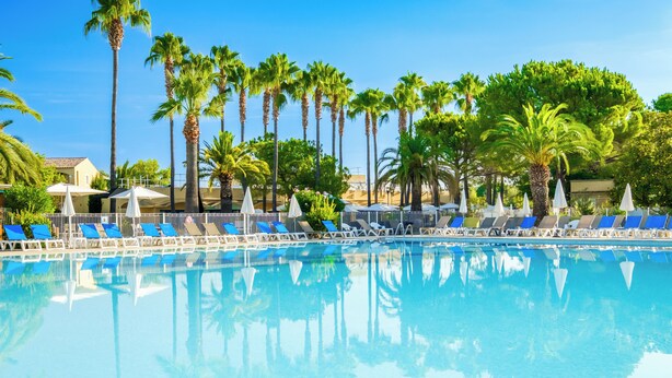 A swimming pool with blue lounge chairs and umbrellas, surrounded by palm trees. - Pierre & Vacances Residence Cannes Mandelieu (Mandelieu-La-Napoule)