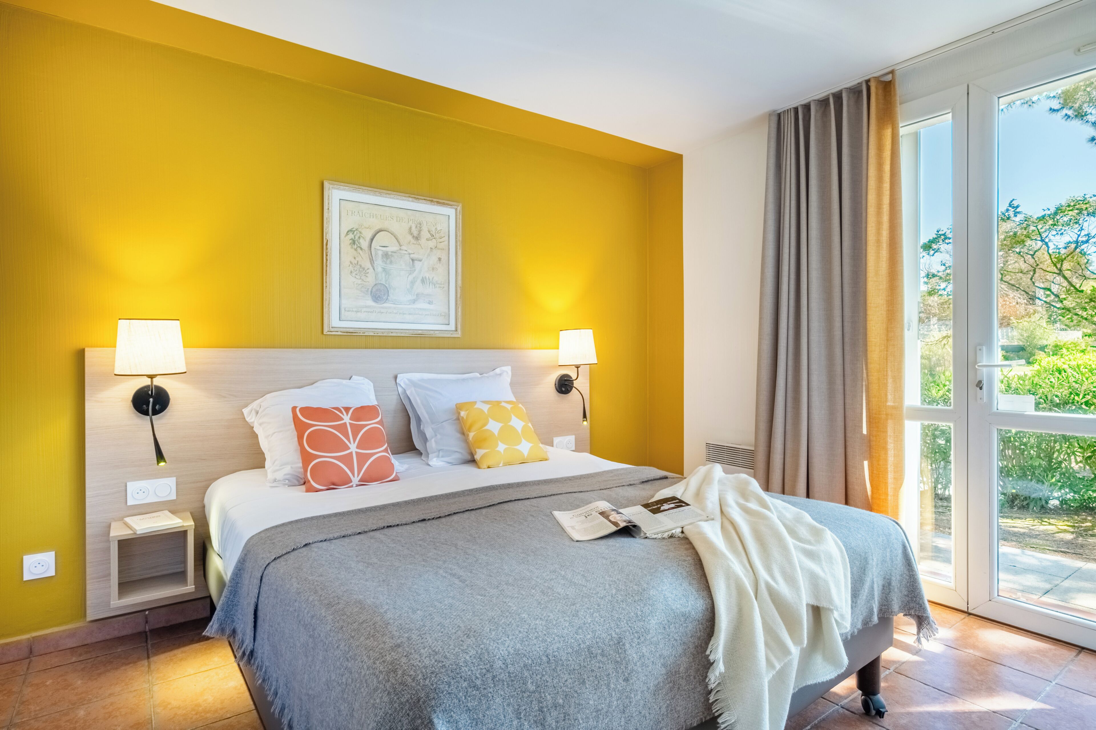A hotel room with a bed, two bedside lamps, a framed picture above the bed, a window with curtains, and a view of greenery outside.