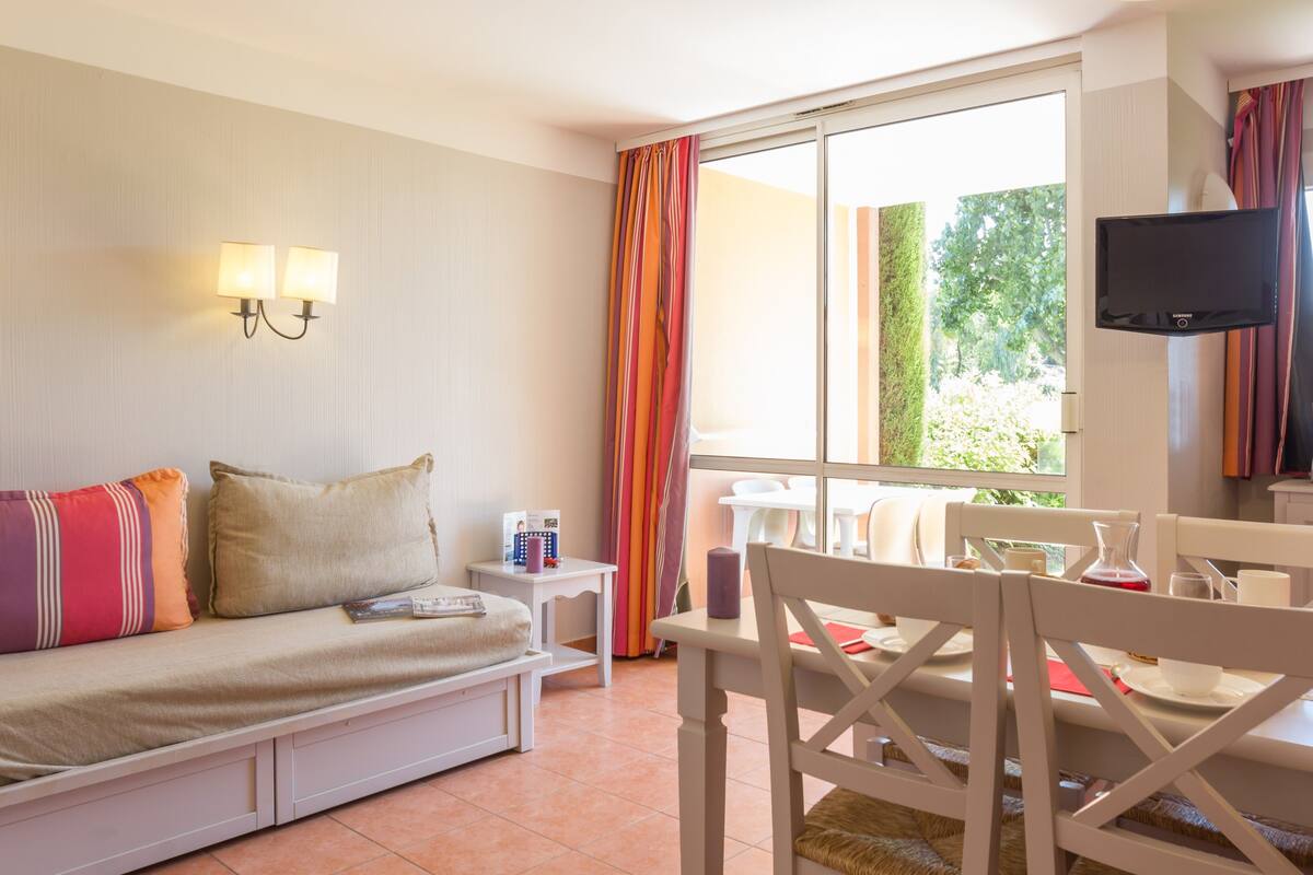 Ein Zimmer mit einem Bett, einem kleinen Tisch mit Stuhl, einem an der Wand montierten Fernseher und einem Fenster mit Vorhängen.