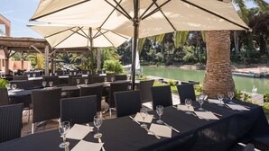 An outdoor dining area with tables set for two, each with a tablecloth, wine glasses, and cutlery. Large umbrellas provide shade, and the setting overlooks a body of water with a palm tree and greenery in the background. - Pierre & Vacances Residence Cannes Mandelieu (Mandelieu-La-Napoule)