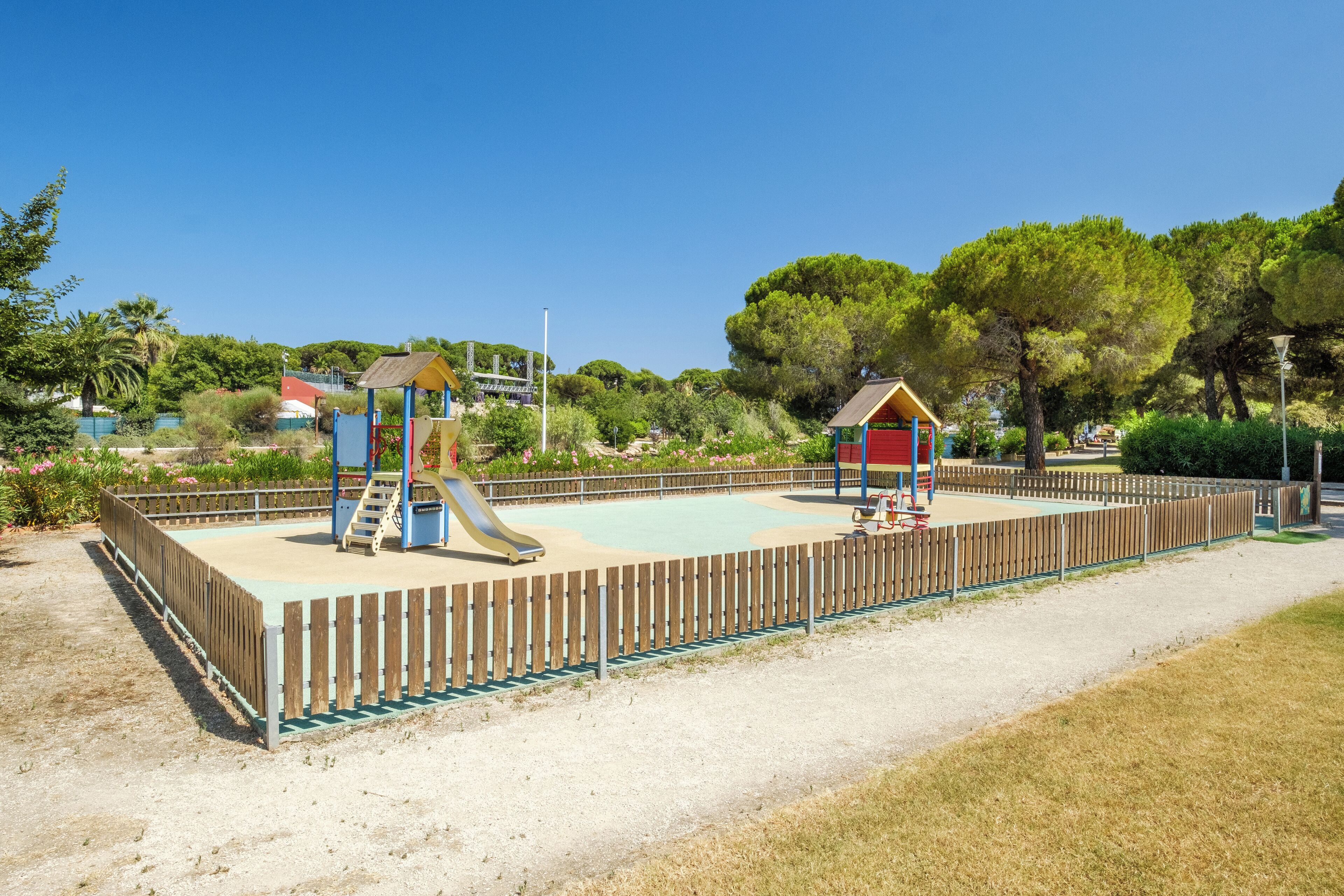 A playground with a slide and climbing structures, surrounded by a wooden fence and trees.