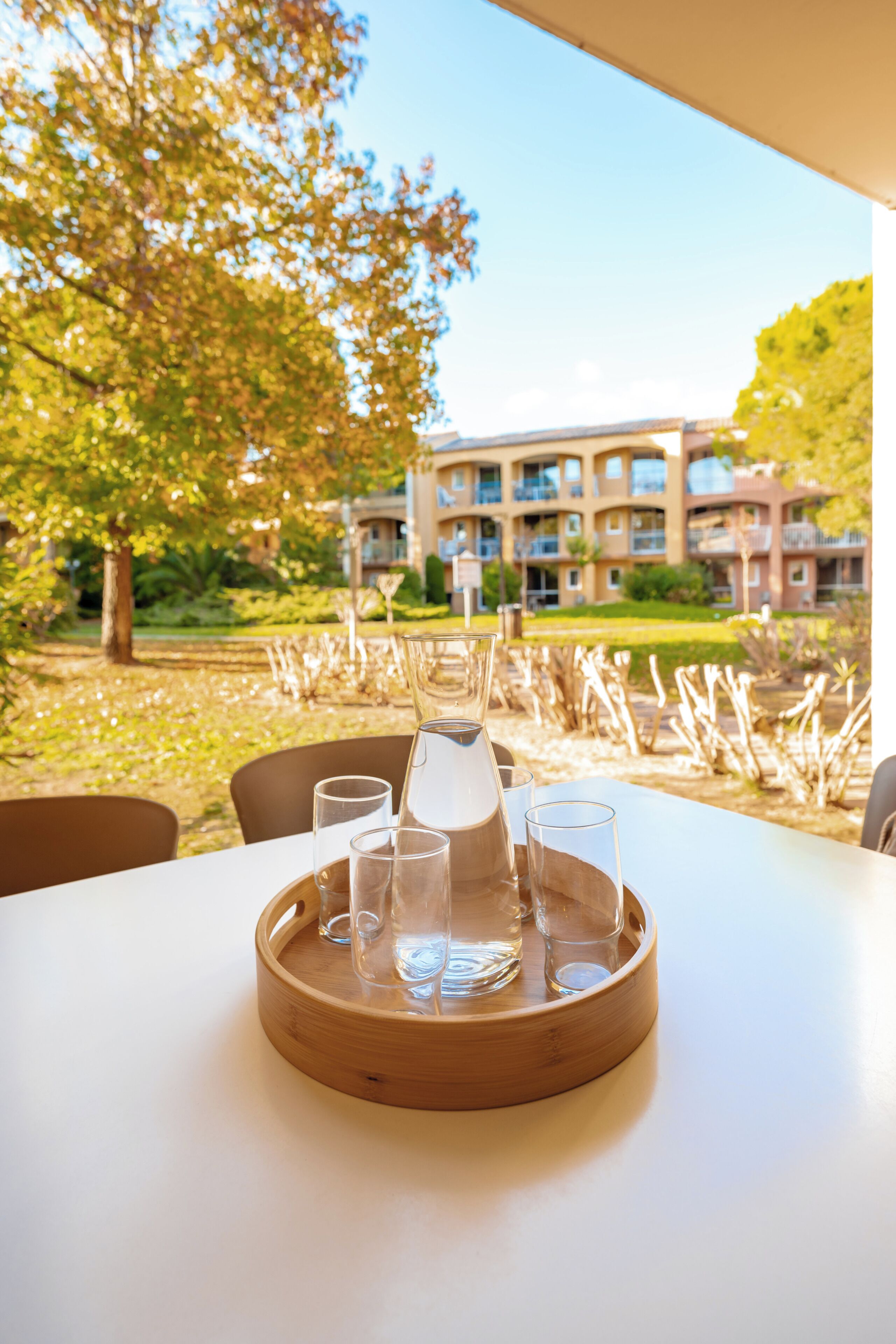 A table with a tray holding four glasses and a bottle, set against a backdrop of a building and trees.