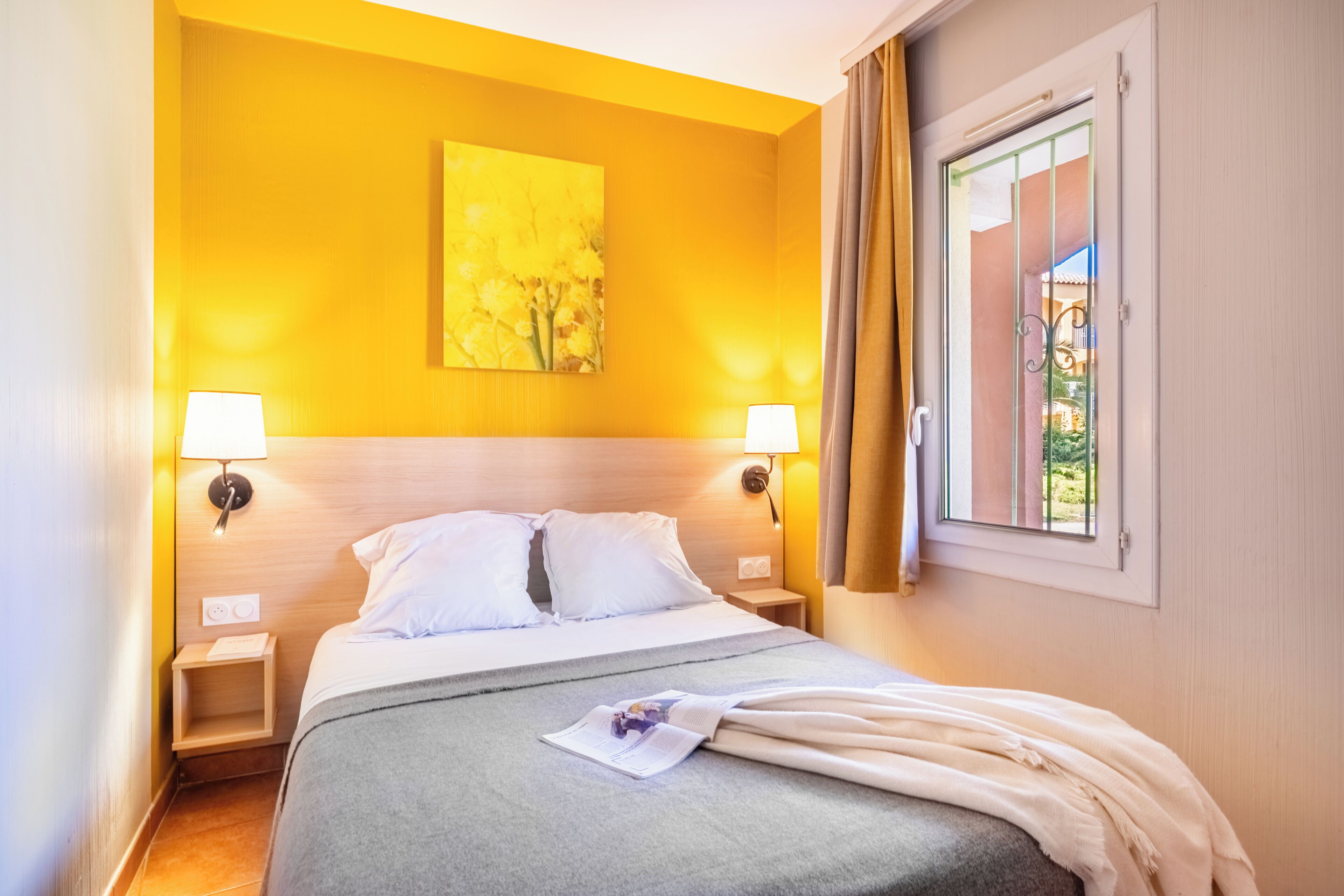 A hotel room with a bed, two bedside lamps, a window with curtains, and a painting on the wall.