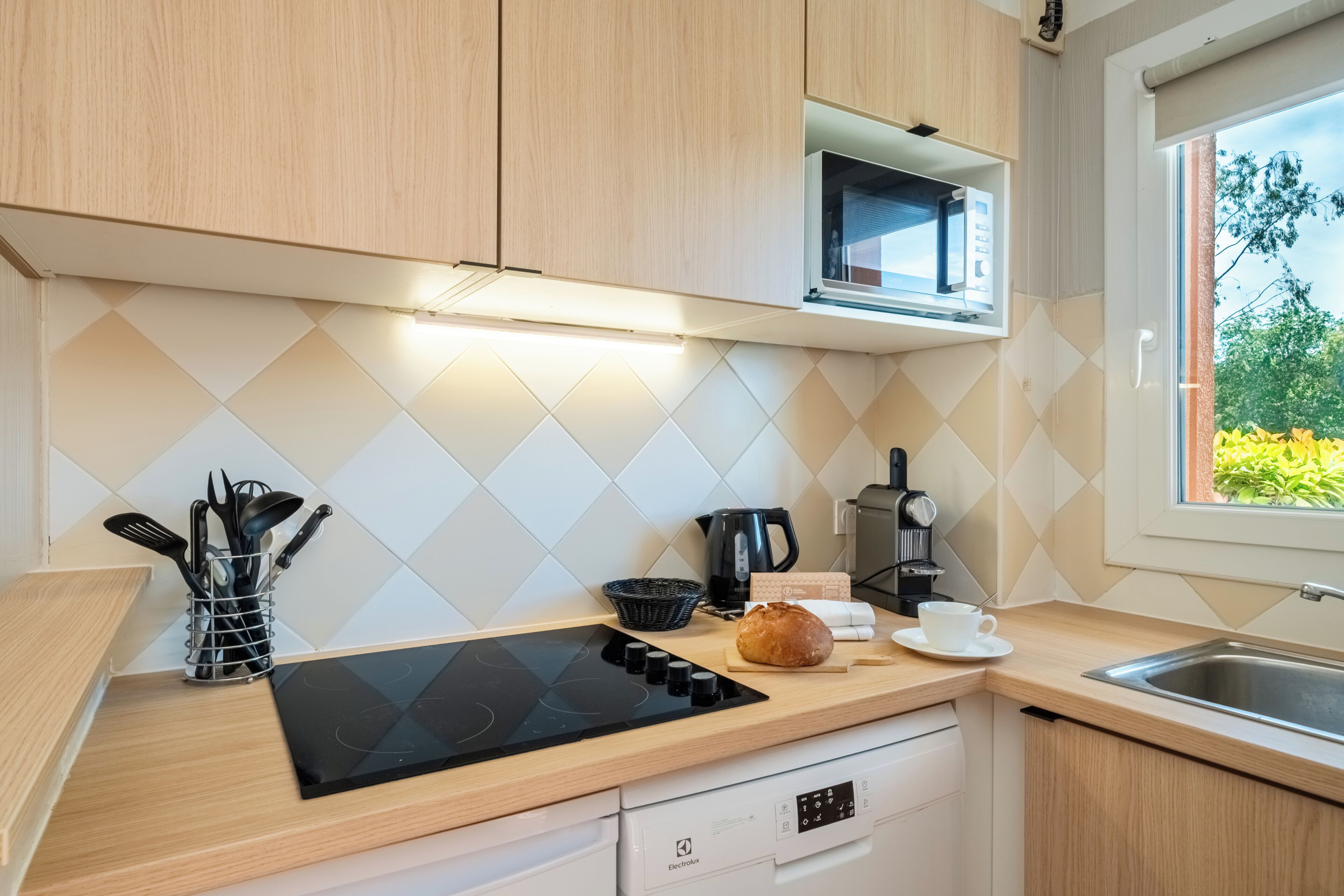 Una cocina moderna con una placa de inducción negra, una cafetera, un pan y una ventana con vistas al verdor.