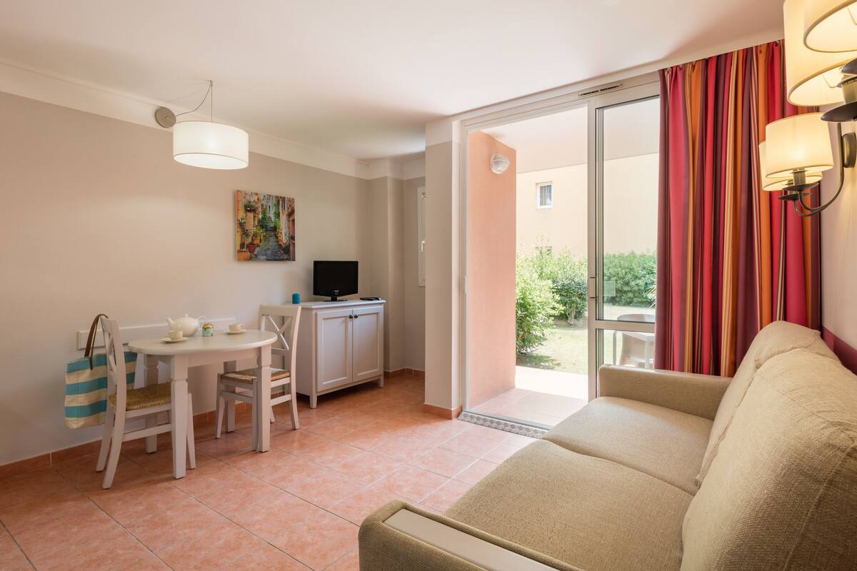 Ein Wohnzimmer mit einer Couch, einem Esstisch mit Stühlen, einem Fernseher und einem Fenster mit Vorhängen.
