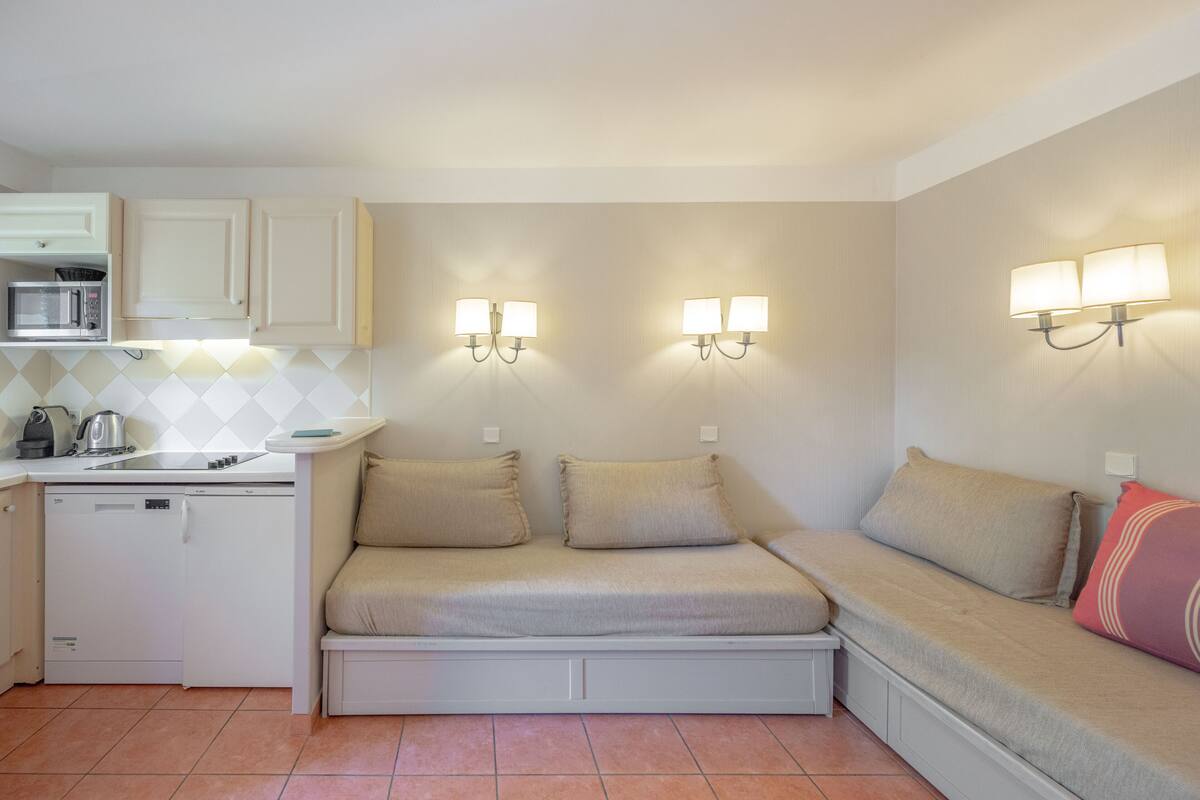Eine kompakte Küchenzeile mit Mikrowelle, Wasserkocher und Geschirrspüler. Zwei Wandleuchten und ein Sofa mit Kissen sind vorhanden.