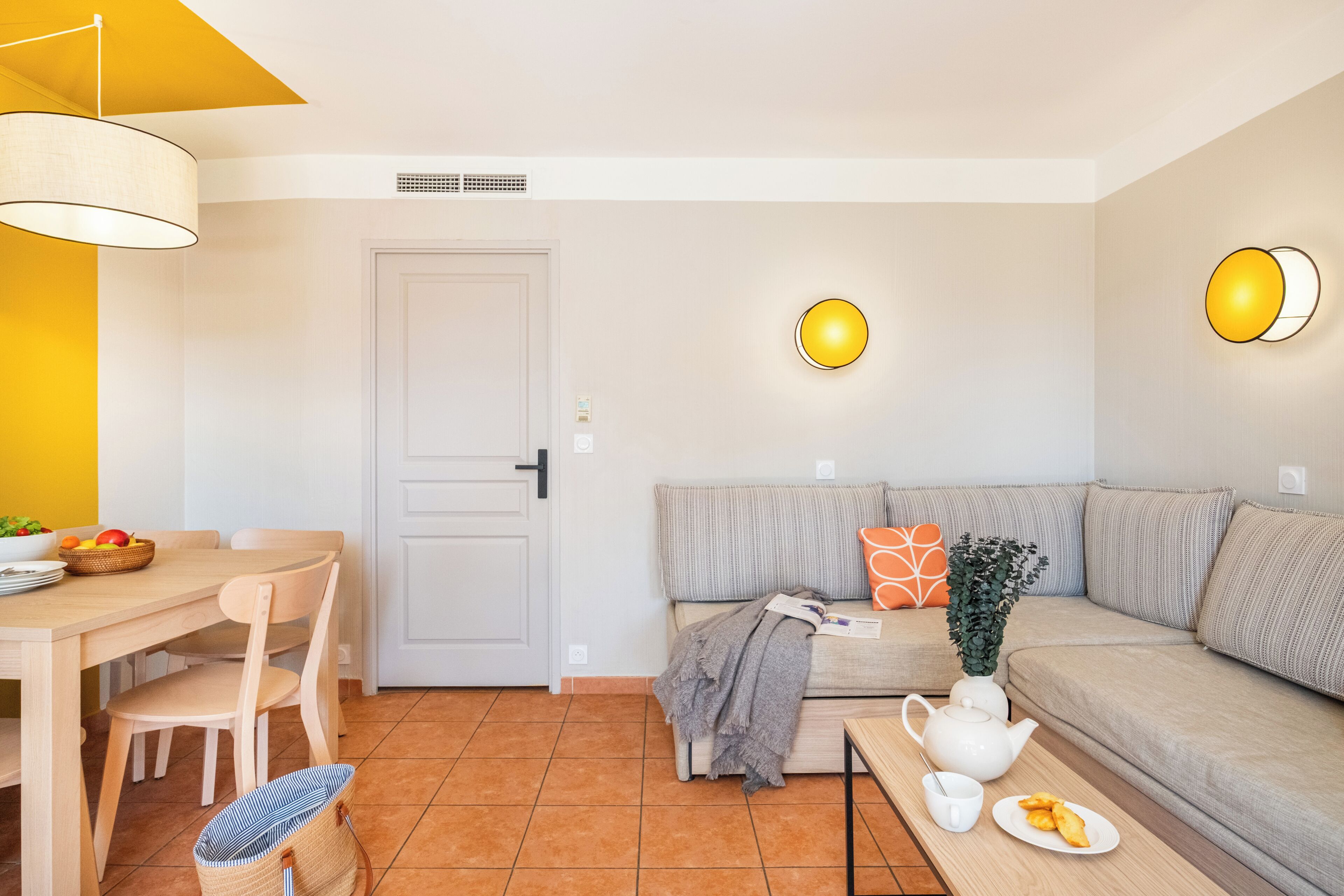 A modern living room with a sofa, a dining table with chairs, a coffee table with a teapot, and a basket of fruit.