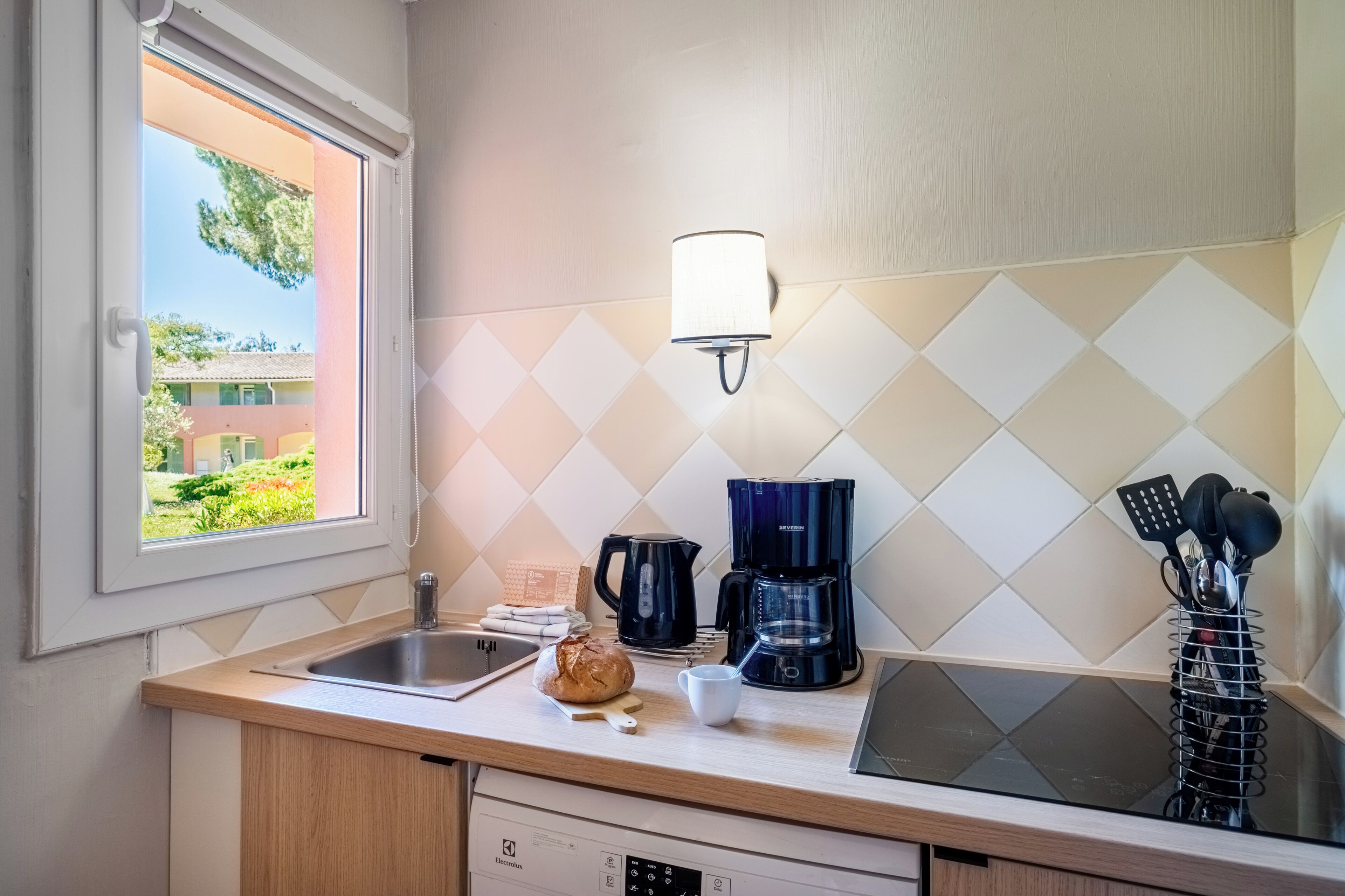 A kitchen with a window, a coffee maker, a kettle, a toaster, and a sink.