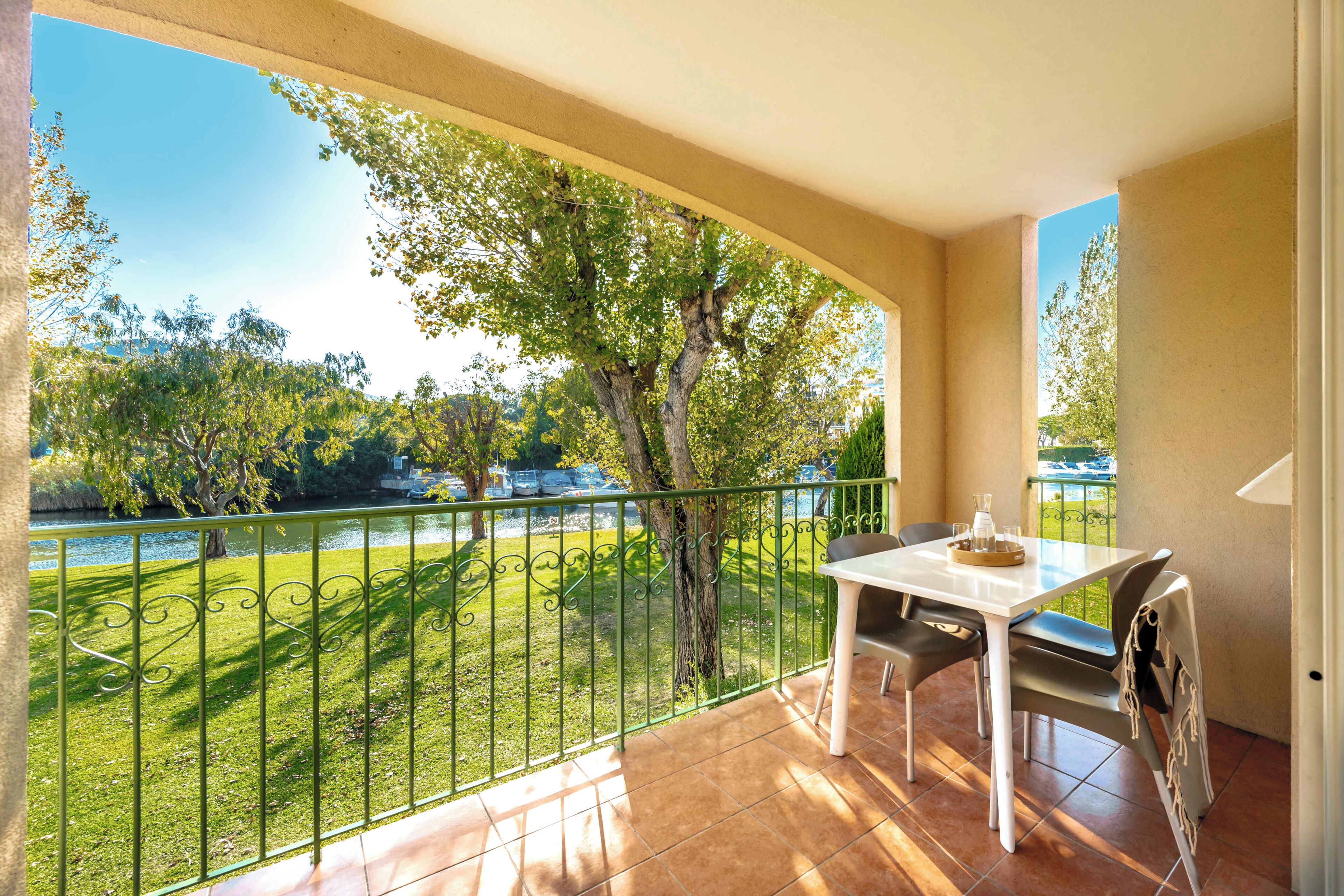 A balcony with a table and chairs, a view of a park with a tree, and a railing.