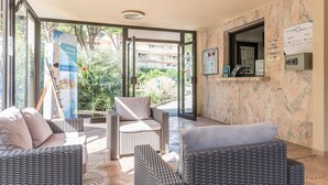 A hotel lobby with wicker furniture, a glass door leading outside, and a weather information board. - Pierre & Vacances Résidence Les Jardins Ombragés (Mandelieu-La-Napoule)