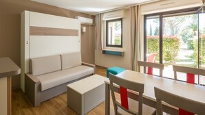 A modern, compact room with a sofa, a small table, and a window providing a view of greenery outside. - Pierre & Vacances Résidence Les Jardins Ombragés (Mandelieu-La-Napoule)