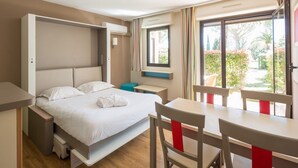 A modern hotel room with a bed, a desk, and a chair. There is a window with curtains and a view of greenery outside. - Pierre & Vacances Résidence Les Jardins Ombragés (Mandelieu-La-Napoule)
