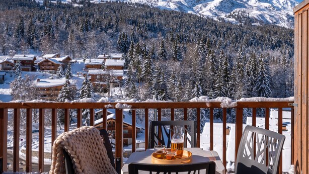 A wooden deck with a table and chairs, overlooking a snowy mountain landscape with cabins. - Résidence Pierre & Vacances Les Ravines (Les Allues)