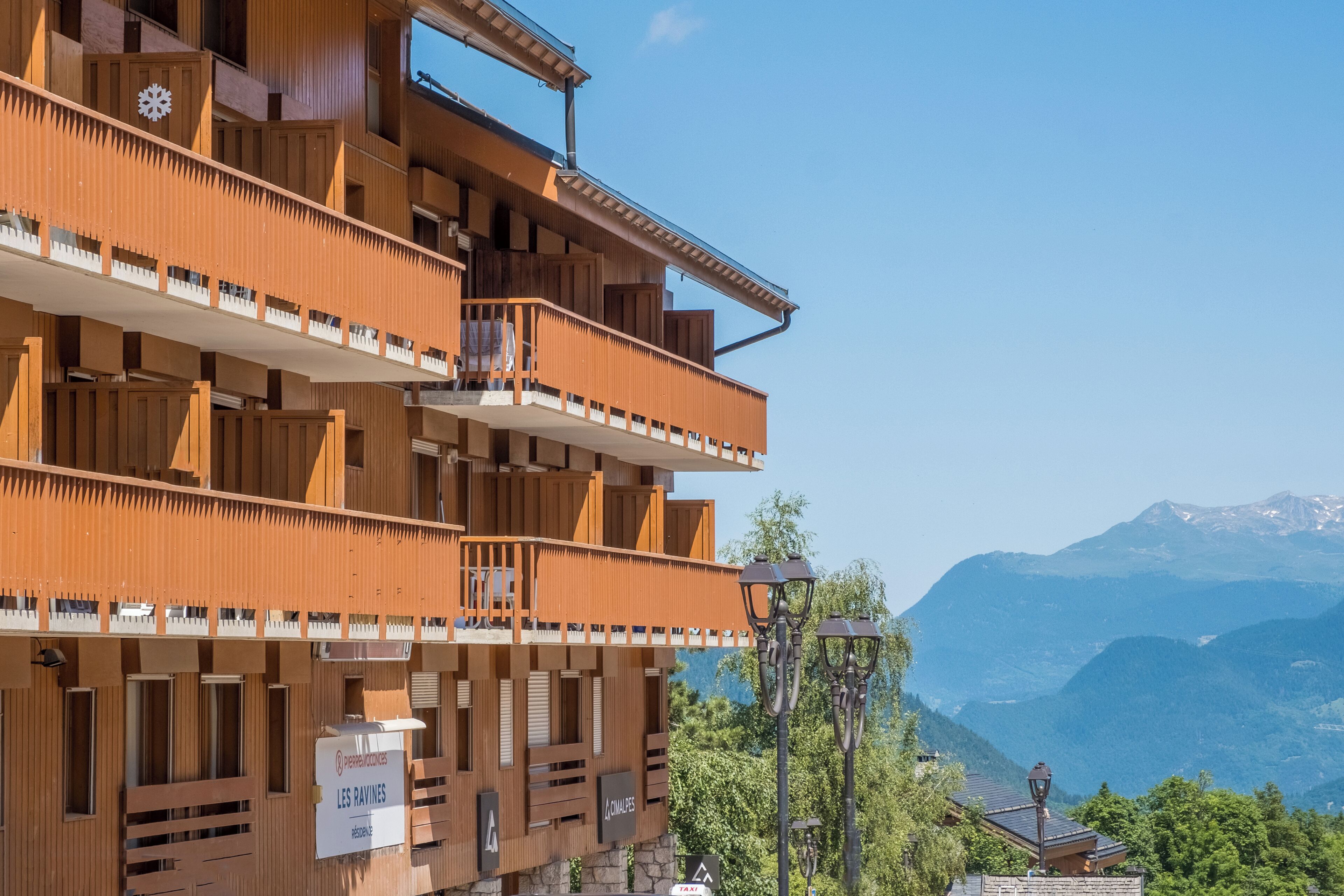 A multi-story wooden building with balconies, a sign indicating 'LES RAVINES,' and a view of mountains in the background.