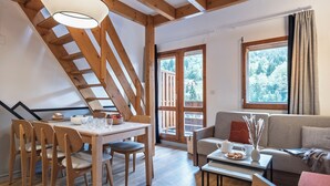 A cozy living area with a wooden staircase, a dining table with chairs, a sofa, and a window with a view of trees. - Résidence Pierre & Vacances Les Ravines (Les Allues)