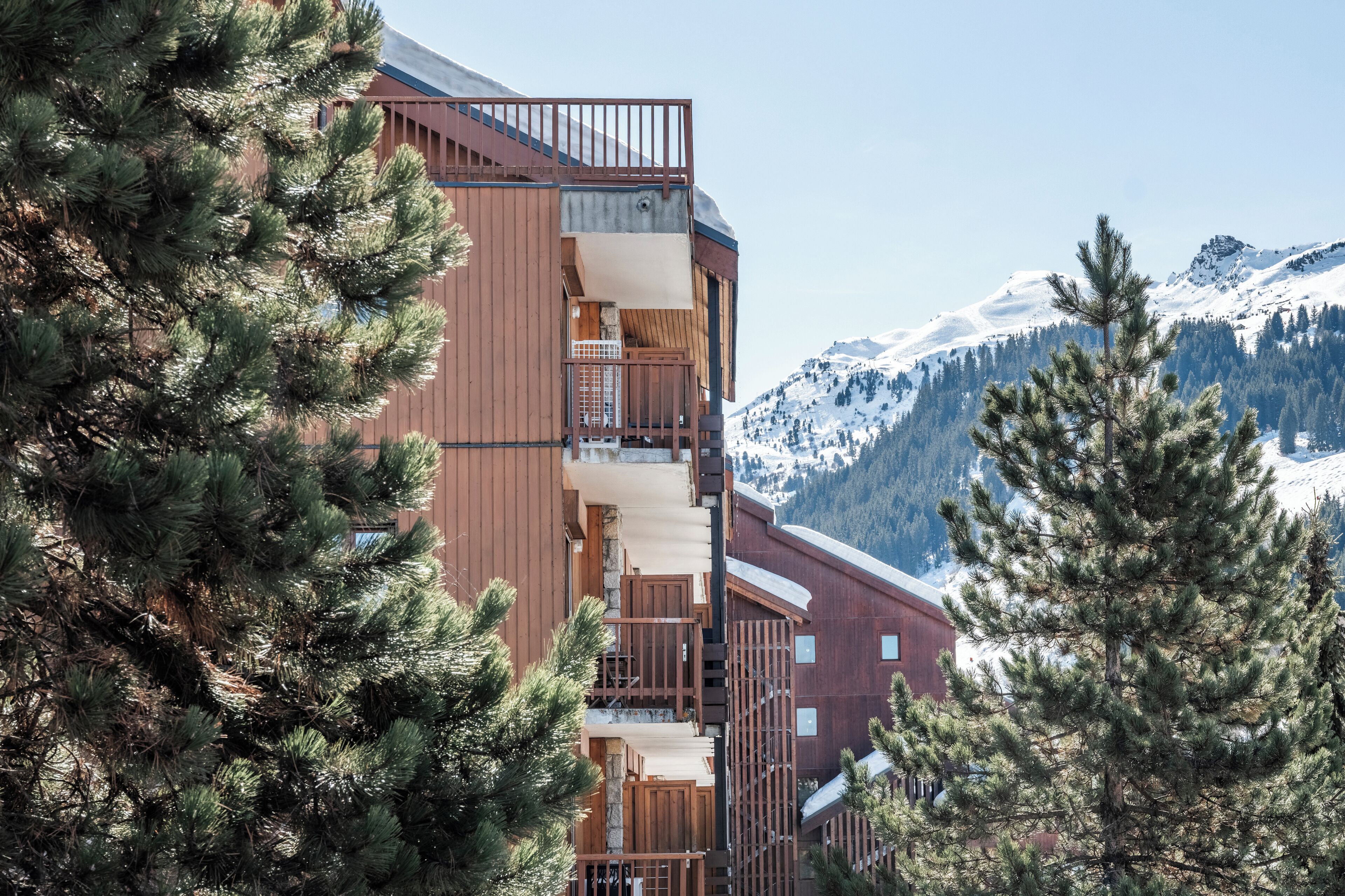 Un edificio de varios pisos con balcones, rodeado de pinos y montañas nevadas al fondo.